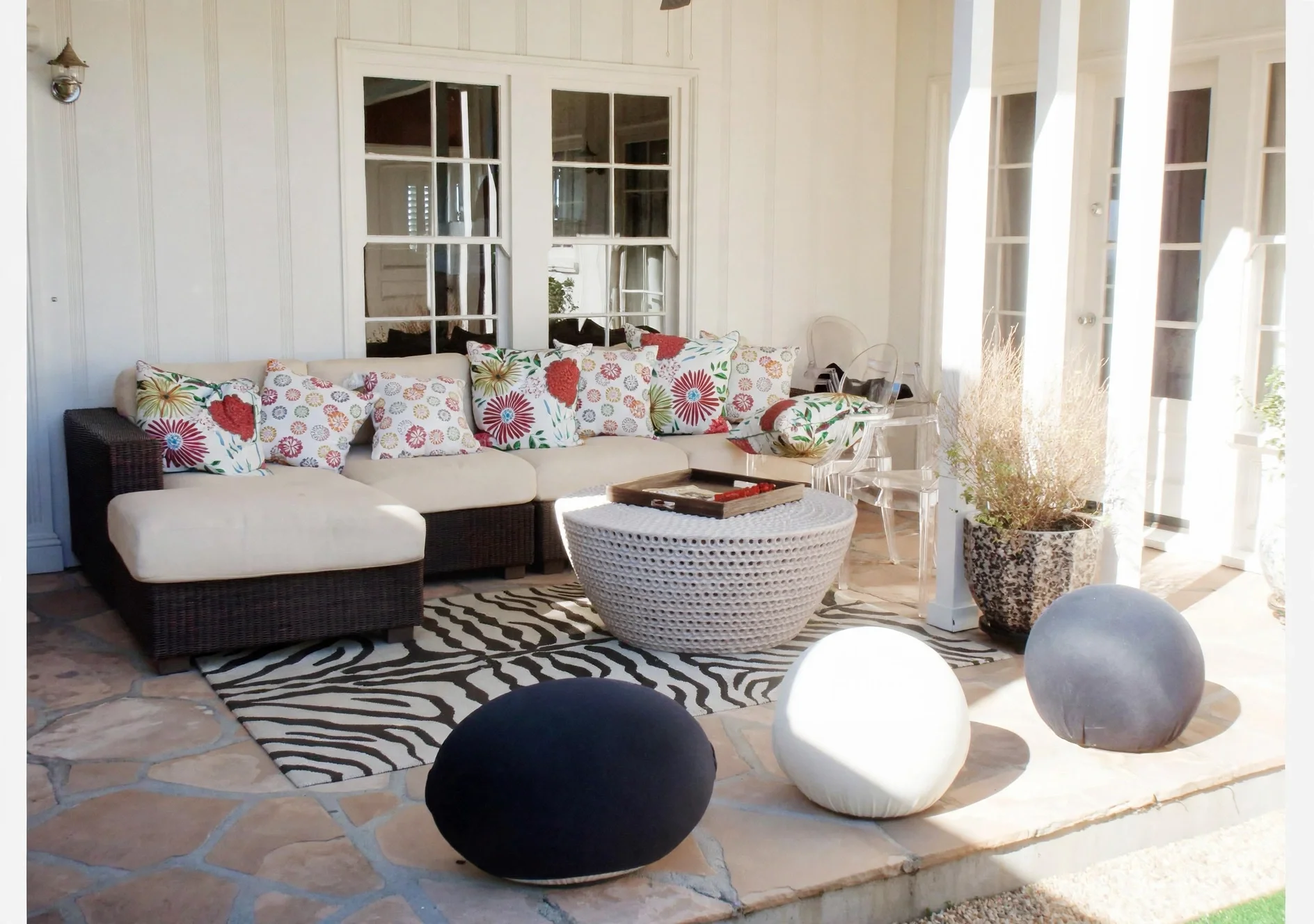 A cozy living room with a sectional sofa adorned with floral-patterned pillows, a round wicker coffee table, zebra print rug, and large windows with white curtains allowing sunlight to brighten the space. There are decorative elements including a potted plant and hanging light fixtures.