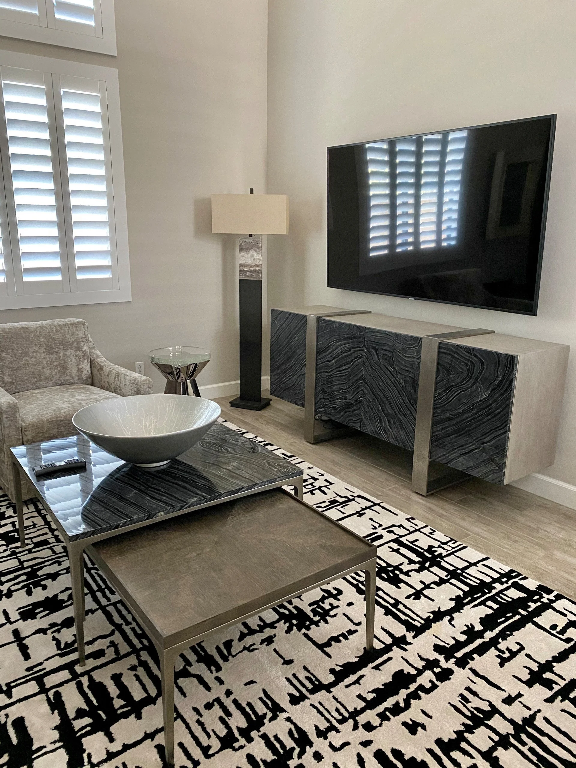 Living room with a modern television mounted on the wall, a marble and metal console table, a patterned black and white rug, a beige sofa, a glass-topped coffee table, a standing lamp, and window blinds.