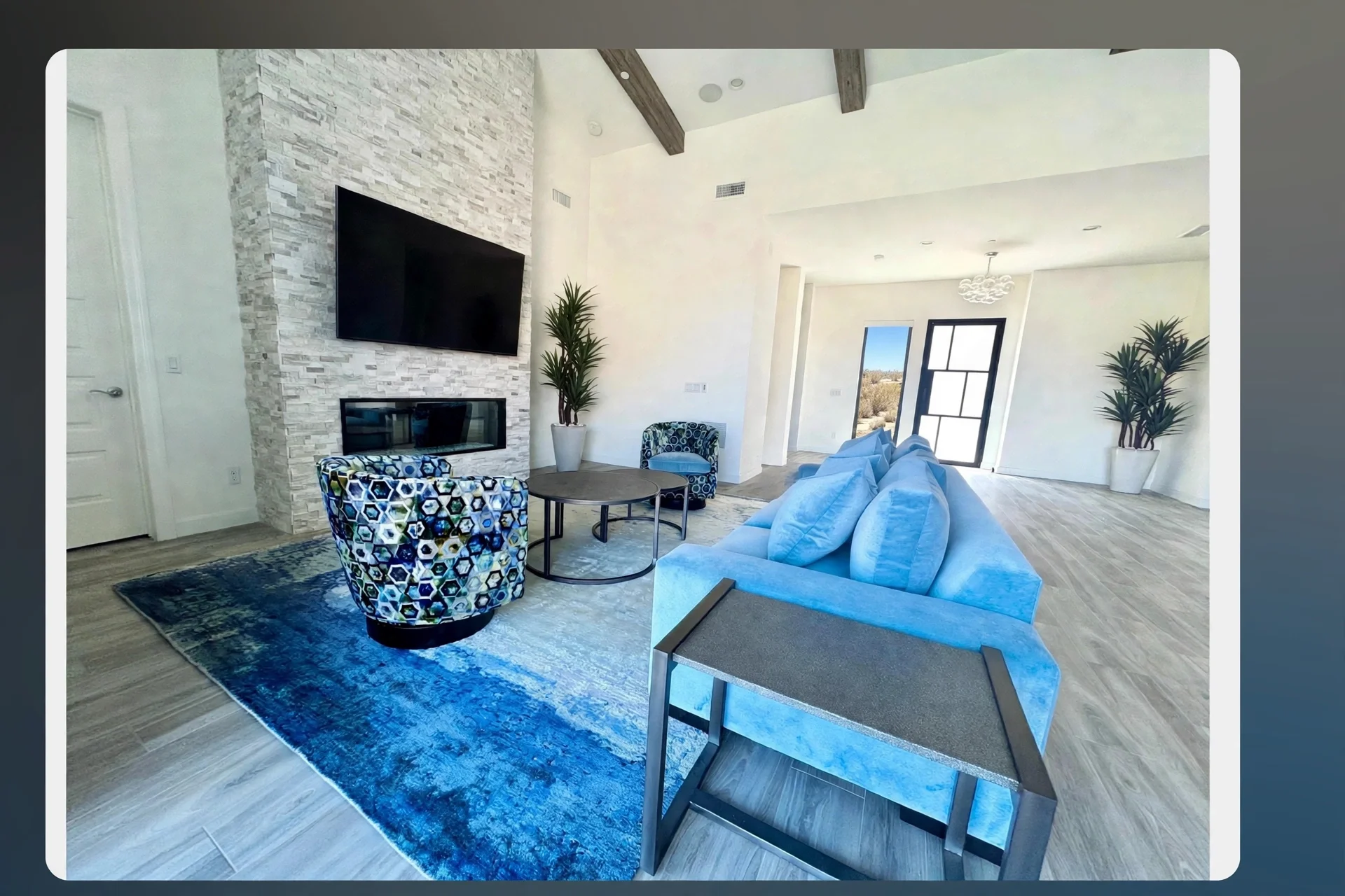 Modern living room with white walls, a stone accent wall featuring a mounted flat-screen TV and a fireplace, a blue sofa, a patterned armchair, a round coffee table, and large windows with black frames letting in natural light. There are two tall potted plants and a blue area rug.