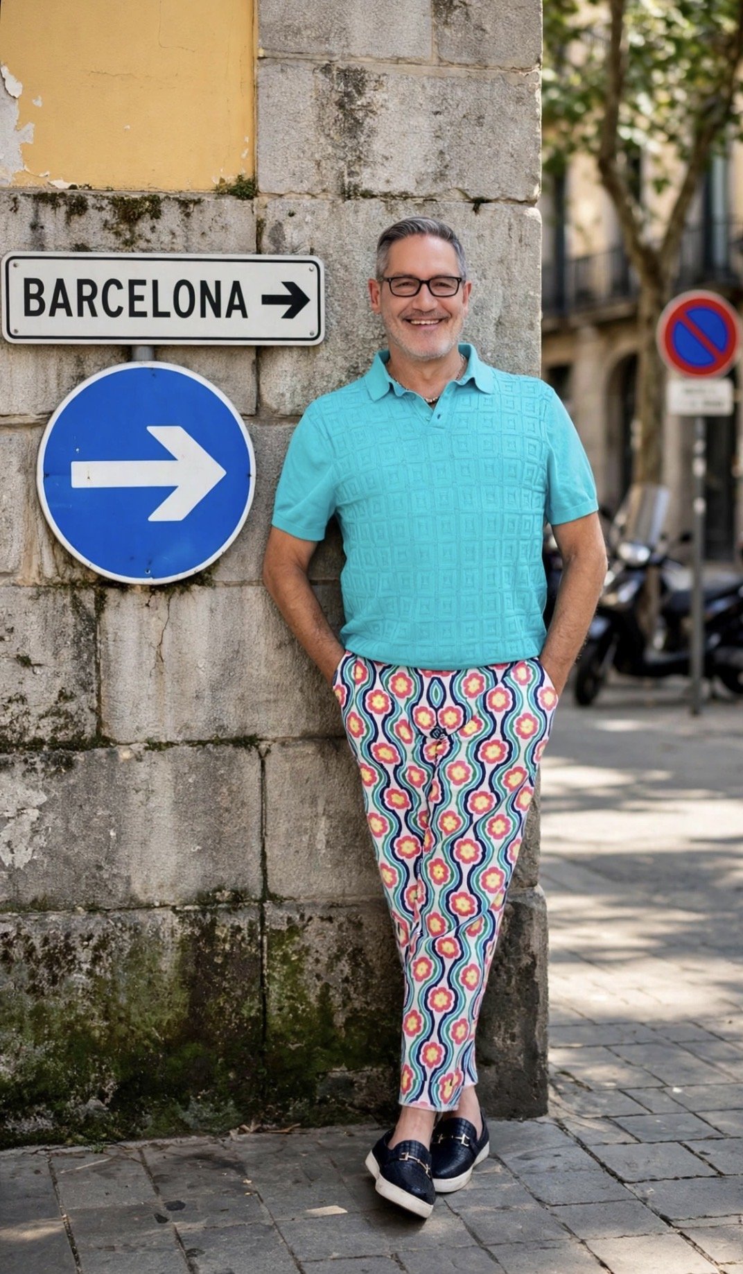A man smiling and standing against a stone wall on a city street, wearing a turquoise shirt, colorful patterned pants, and loafers. There are street signs indicating directions to Barcelona and a blue arrow pointing right.