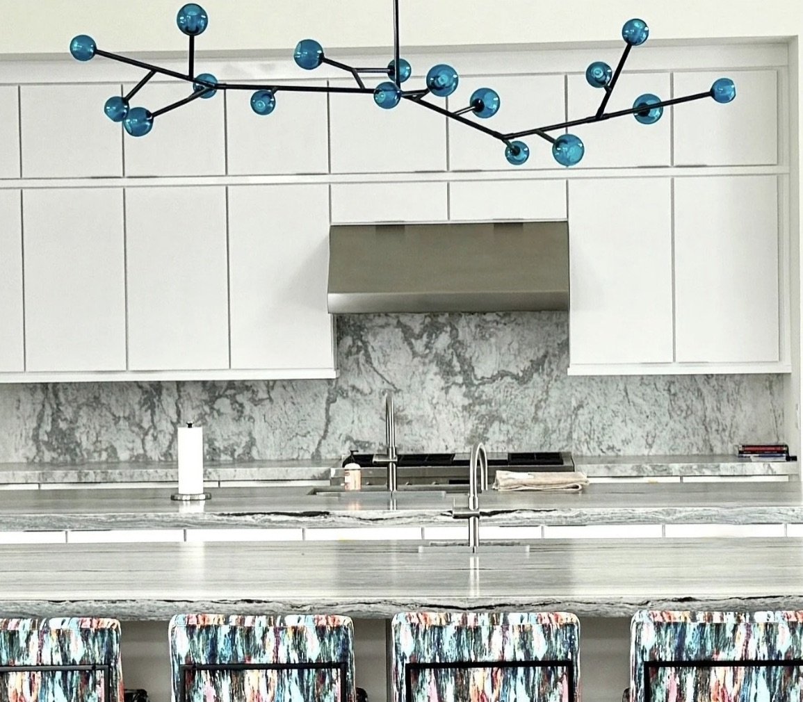 Modern kitchen with white cabinets, marble backsplash, stainless steel range hood, and colorful patterned bar stools at a marble countertop, with a blue geometric chandelier hanging above.