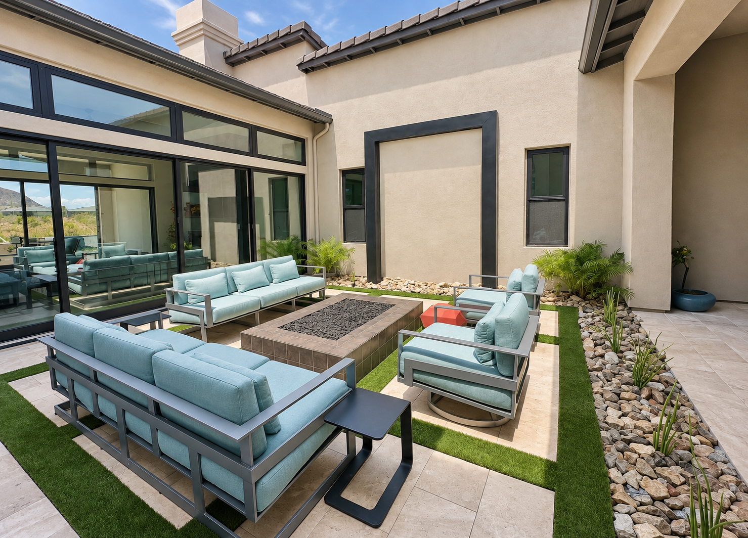 An outdoor patio area with modern furniture, including light blue sofas, armchairs, and black side tables, surrounded by rocks and potted plants, with a glass wall and a house with a chimney in the background.