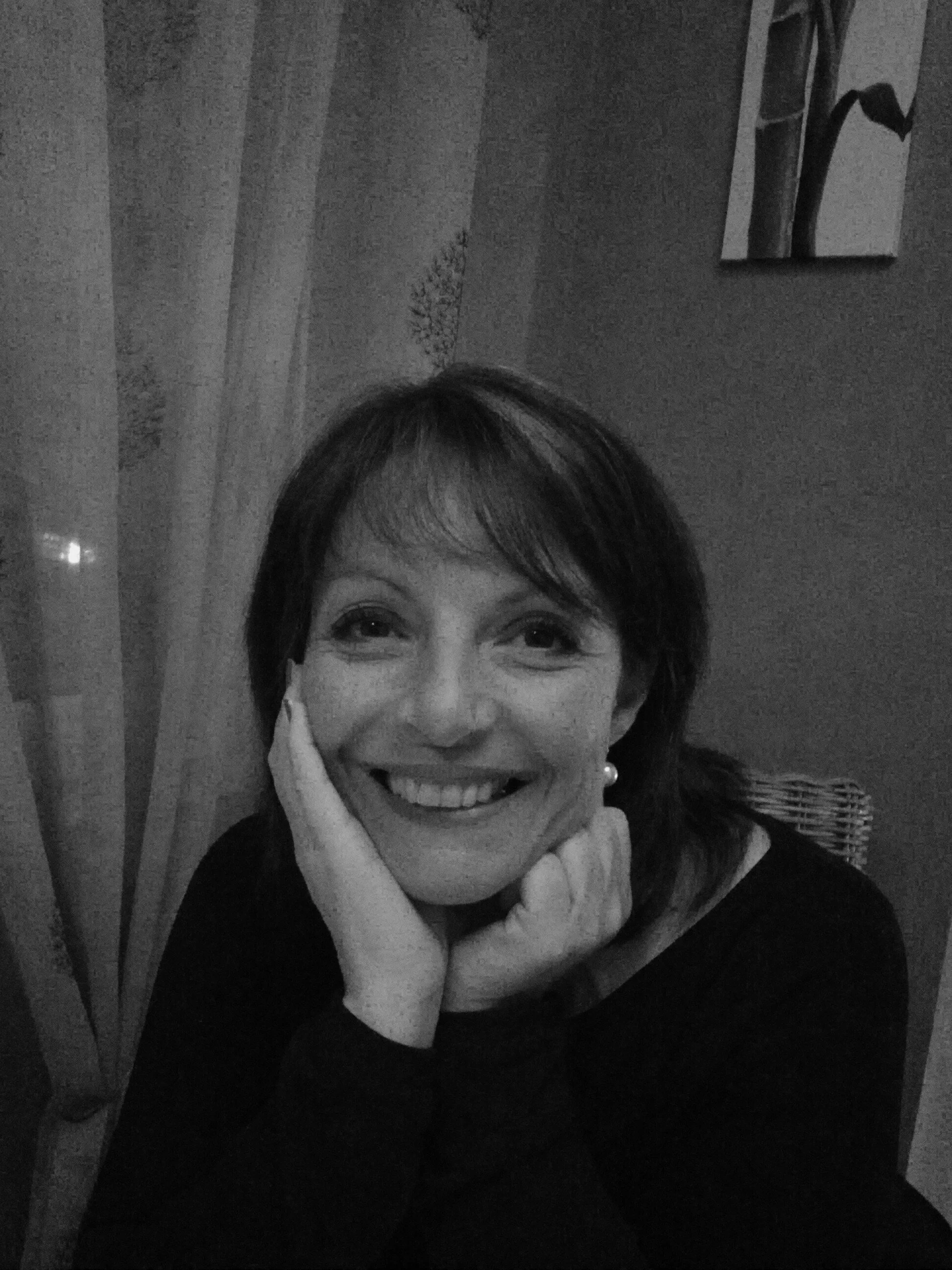 Une femme souriante avec des cheveux courts, portant un pull noir et une écharpe, pense et pose avec ses mains sous le visage, dans une ambiance intérieure en noir et blanc.