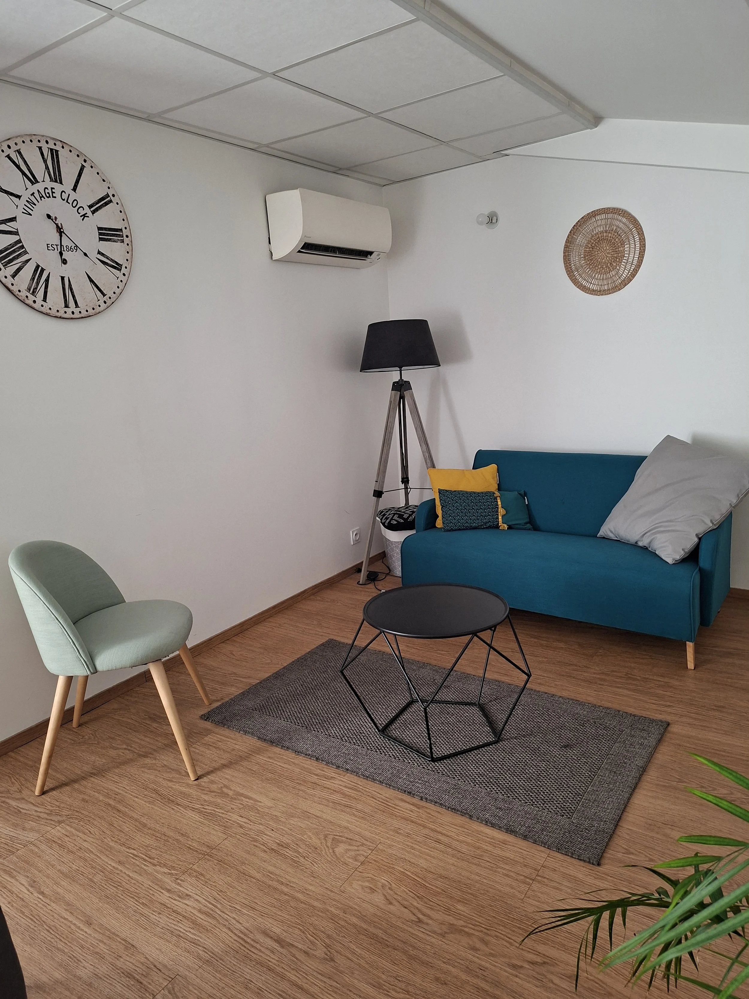 Un salon avec une chaise vert clair, un canapé bleu avec des coussins colorés, une table d'appoint noire, un tapis gris, une lampe sur un trépied, un mur blanc avec une horloge vintage murale, un climatiseur, et une décoration en bois sur le mur.