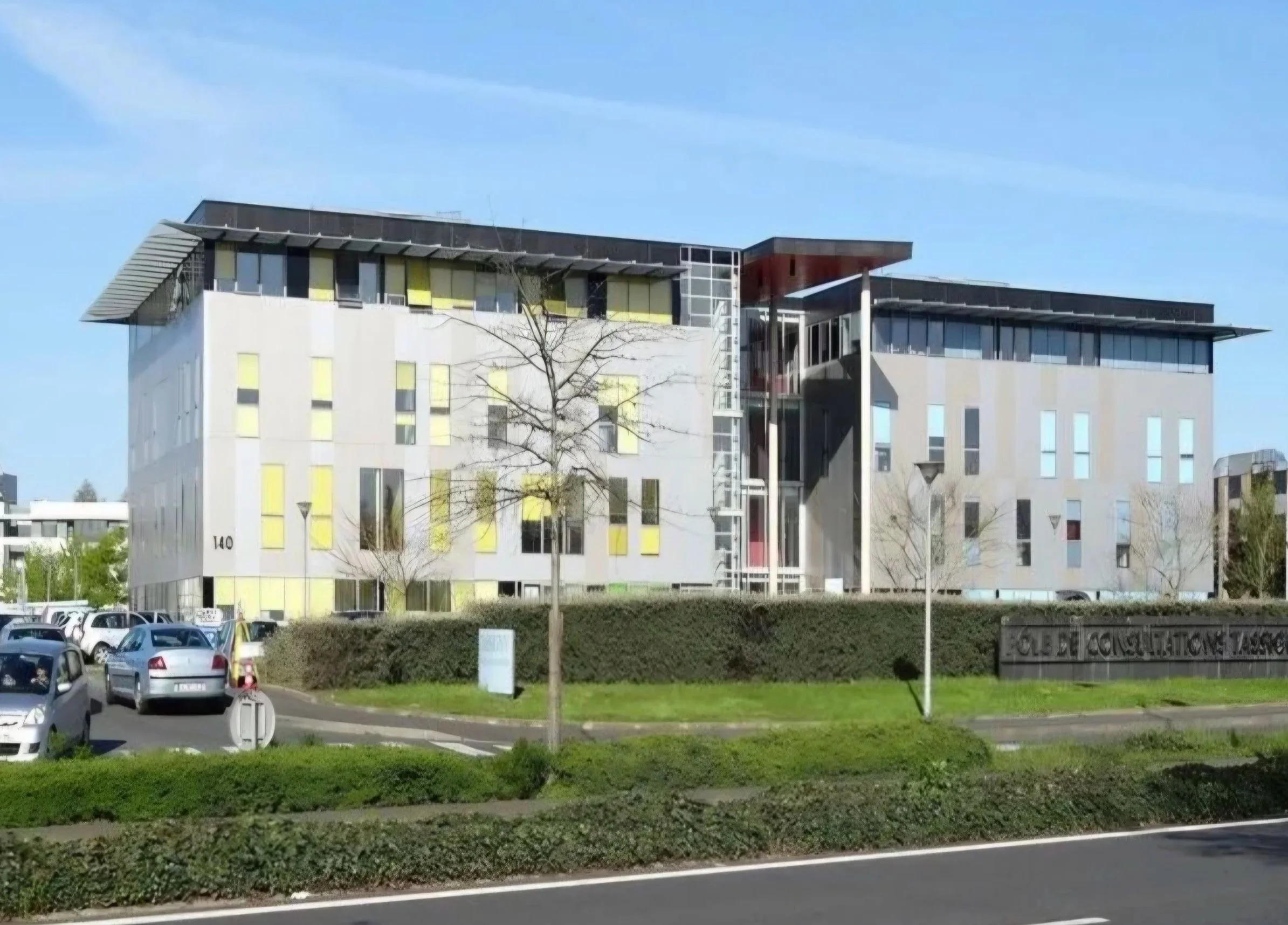 Bâtiment moderne avec plusieurs étages, façades en blanc et accents jaunes, et un toit en partie en porte-à-faux, situé dans un environnement urbain avec des voitures stationnées au premier plan et un panneau d'information.
