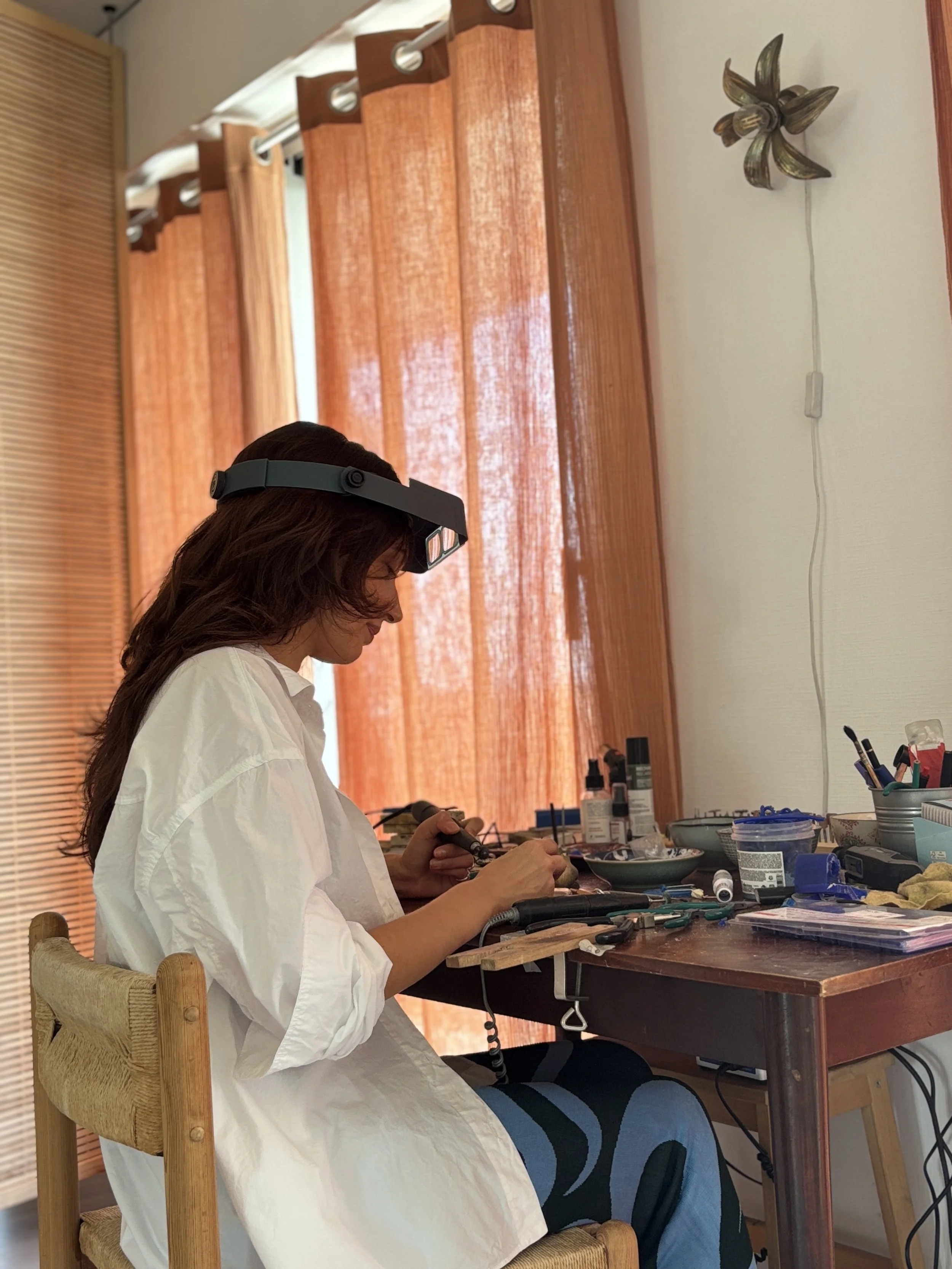 Femme avec un casque de bijoutier, travaillant avec une pièce à main à un établi en bois, entourée de matériel de sertissage, dans un atelier éclairé par la lumière naturelle venant d'une fenêtre avec une lumière plaquée en or.