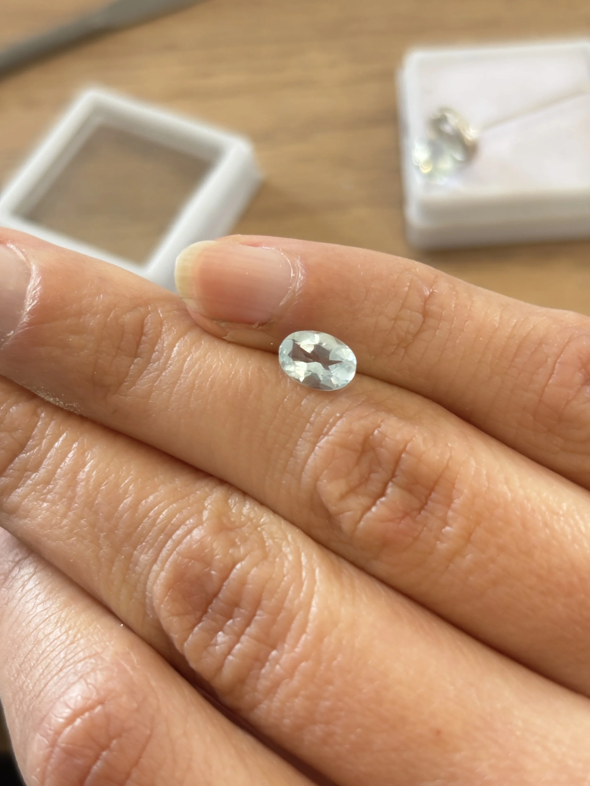 Une aigue-marine taille diamant posée sur le doigt avant d'être façonnée sur une pièce de joaillerie fine. Le savoir-faire artisanal se voit au travers de l'écrin posé sur l'établi dans l'atelier