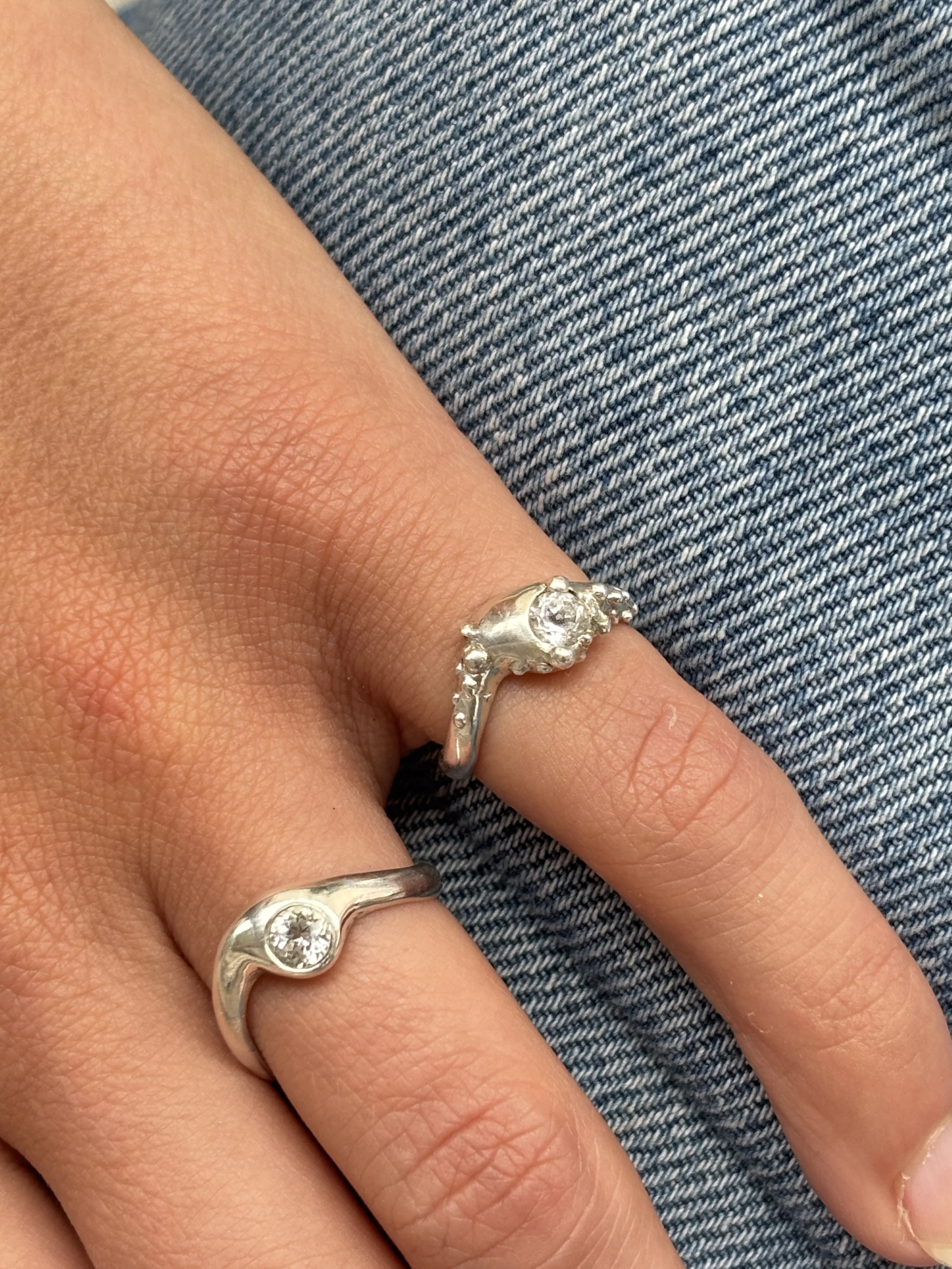 Une main portant deux bagues en argent avec des topazes blanches brillantes, posée sur un tissu en jean pour un style élégant et désirable.