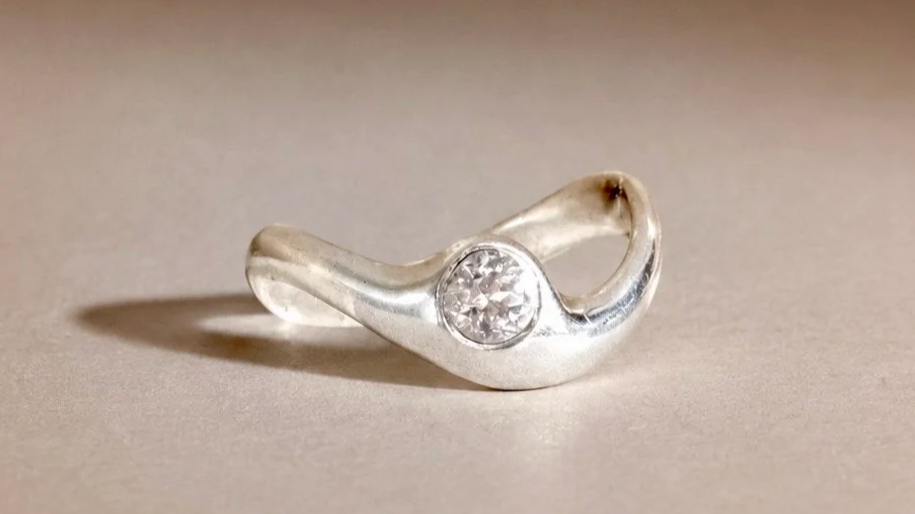 Bague en argent avec un diamant au centre, placée sur une surface claire. Ligne arrondie inspirée par le design futuriste des années 70.