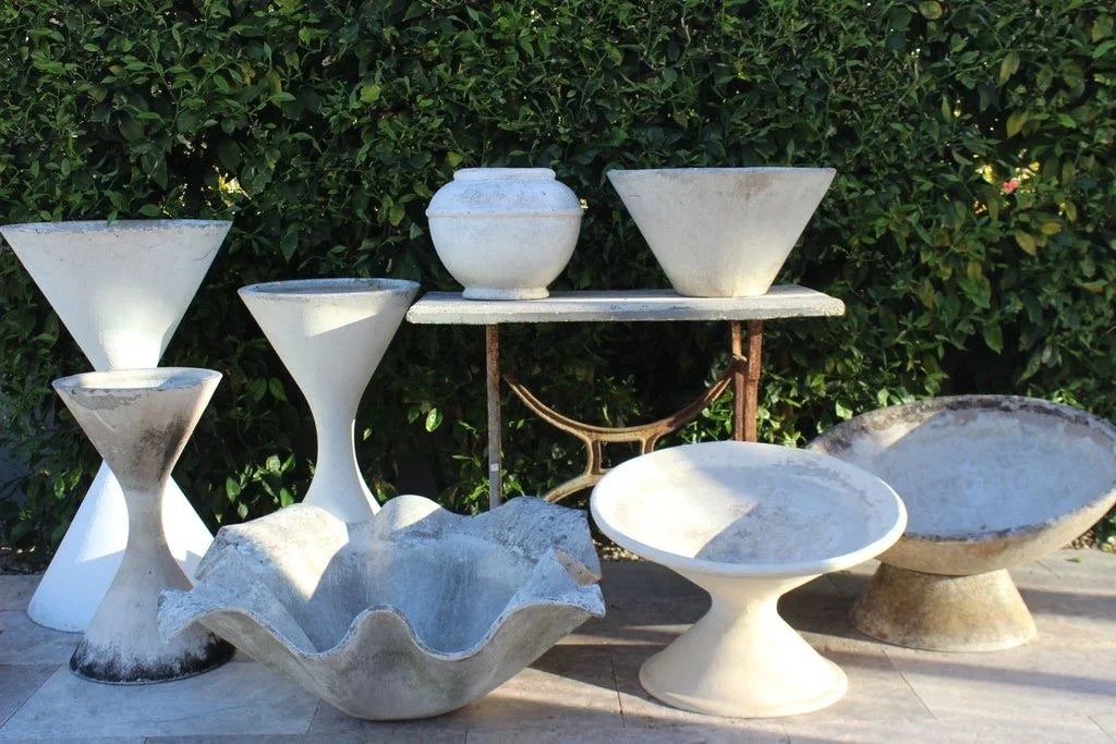 Divers vases et pots en pierre ou en céramique, exposés en extérieur devant un buisson vert. Des formes nveloppantes et organiques rappelant les bagues du site internet.