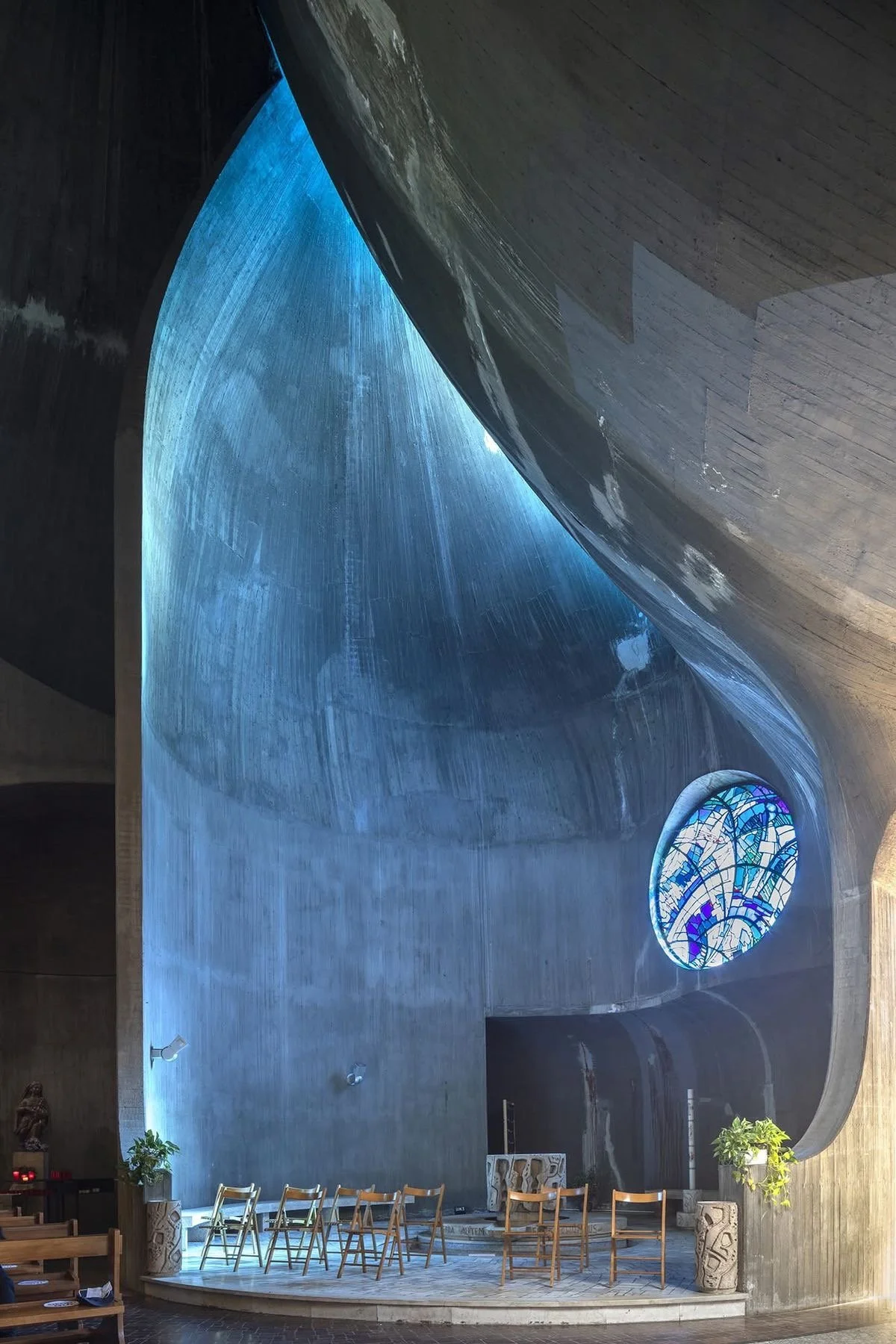 Intérieur d'une chapelle ou d'une église moderne avec un plafond en béton courbé, un vitrail coloré rond et des chaises en bois disposées en cercle