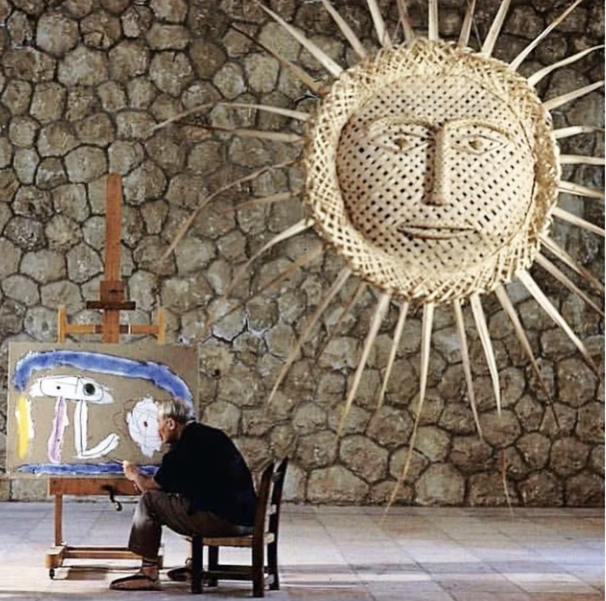 Un homme peint dans un studio avec une œuvre abstraite sur une toile et un grand décor de soleil en osier en forme de visage - soleil accroché au mur en pierre.