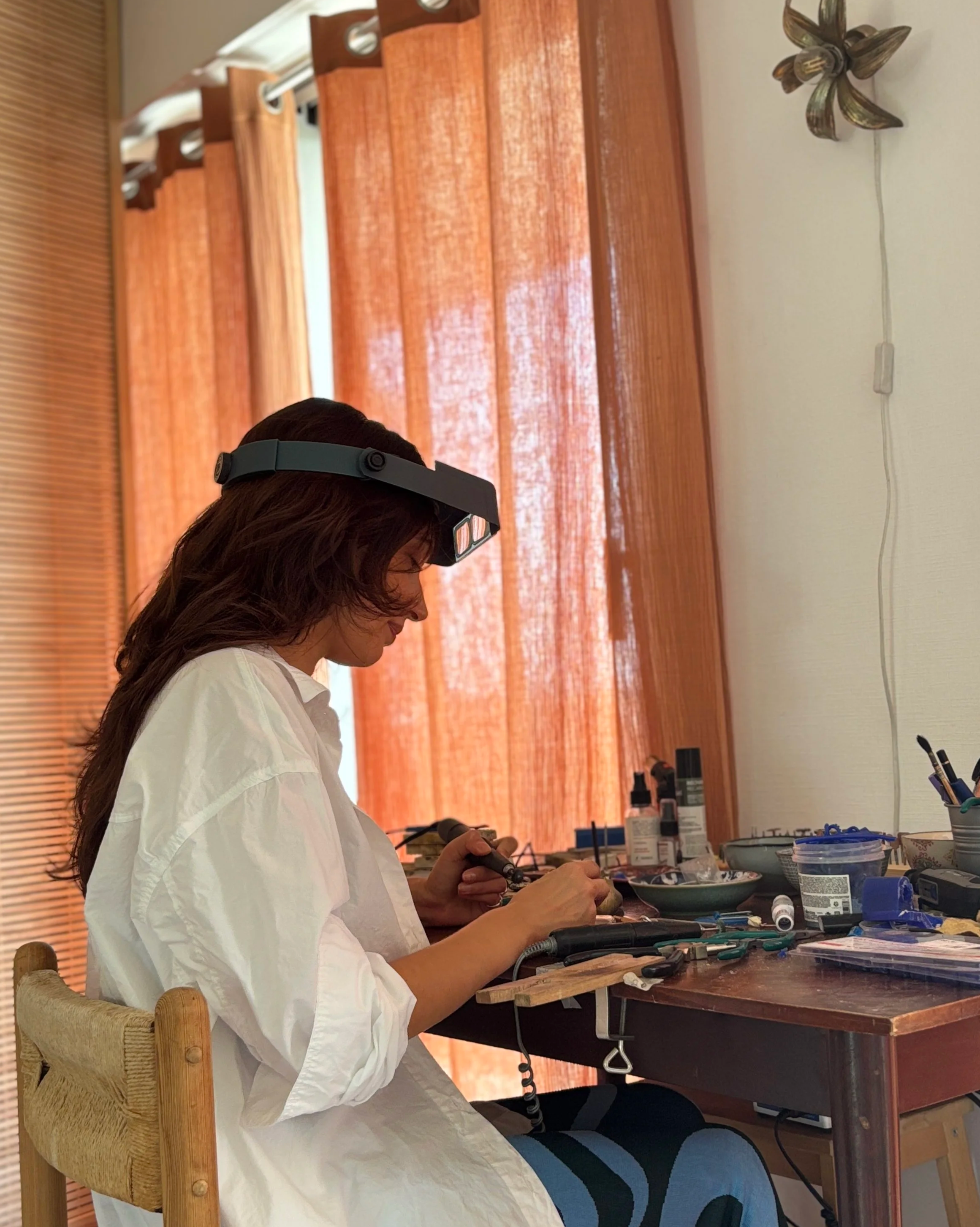 Une femme travaillant à son établi, portant un casque loupe de bijoutier-joaillier, travaillant avec des outils, dans un atelier intérieur, portant une tenue de travail élégante.