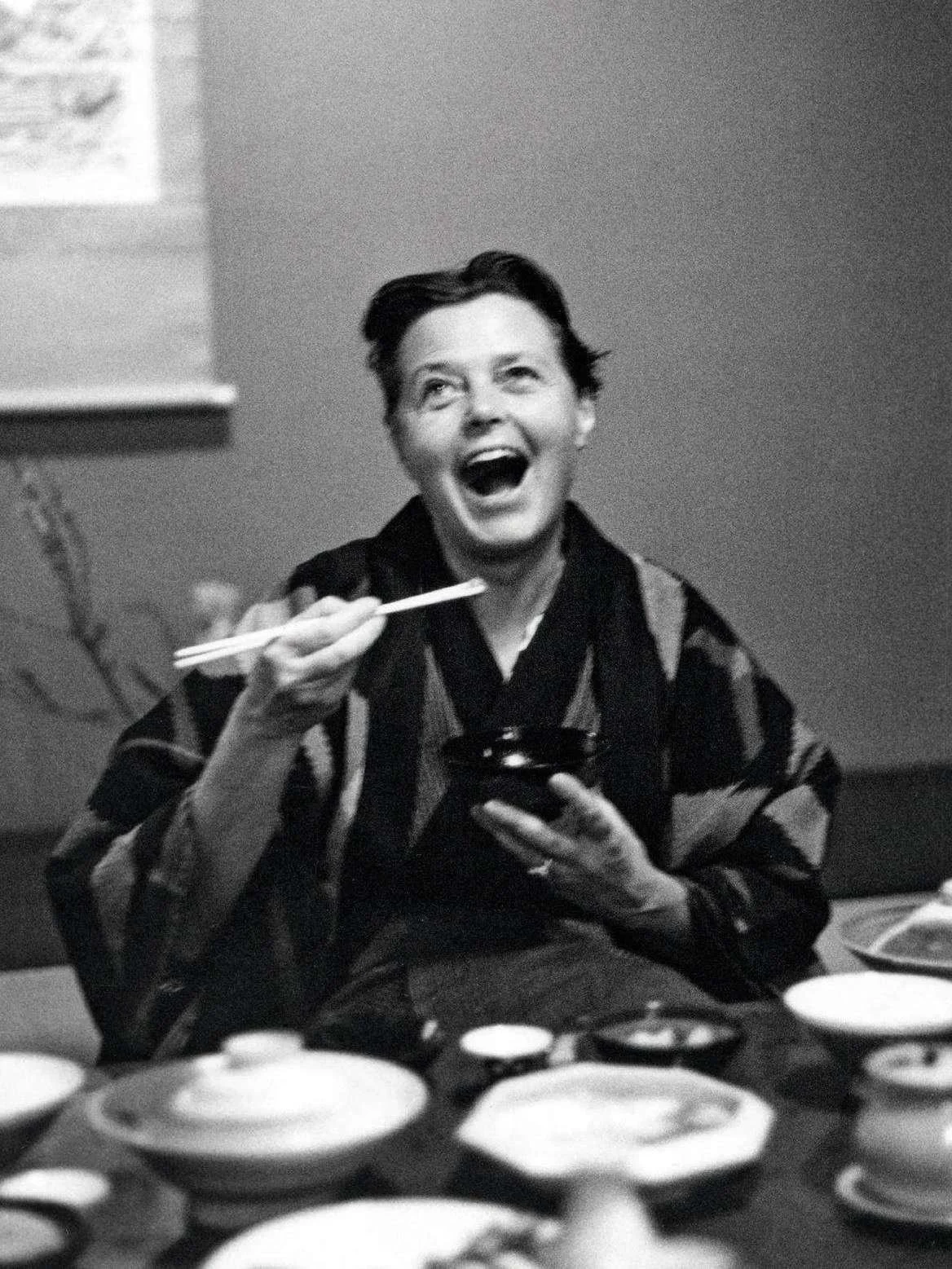 Charlotte Periand souriante, portant un kimono, assise à une table avec plusieurs assiettes, tenant une petite soucoupe et des baguettes, dans une ambiance de repas japonais.