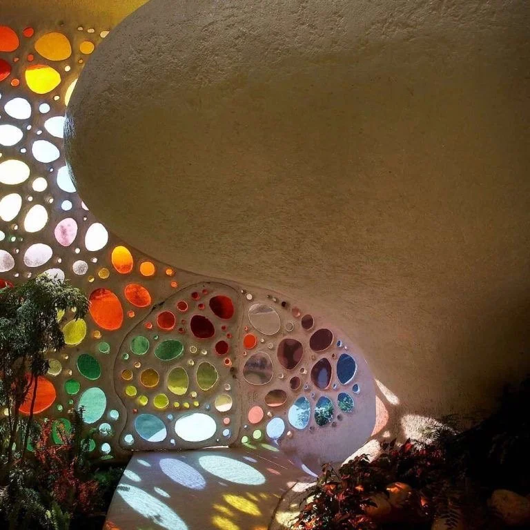 Intérieur d'une structure architecturale avec murs en béton, ornés de nombreux trous de différentes tailles, laissant passer la lumière colorée. Présence de plantes en bas à gauche, ambiance artistique et inventive.