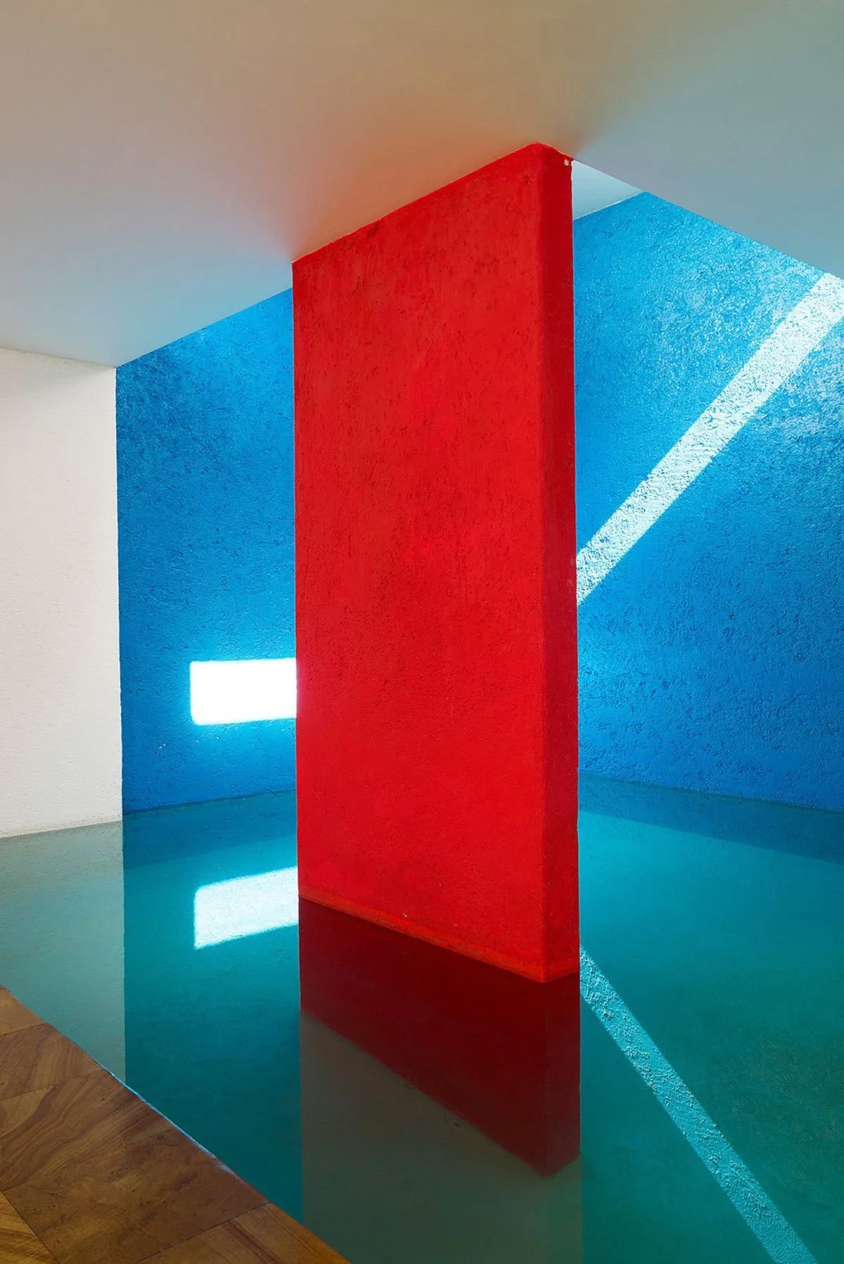 Une installation artistique avec un panneau rouge au centre, entouré de murs bleus et blancs, avec de la lumière naturelle créant des ombres et reflets.