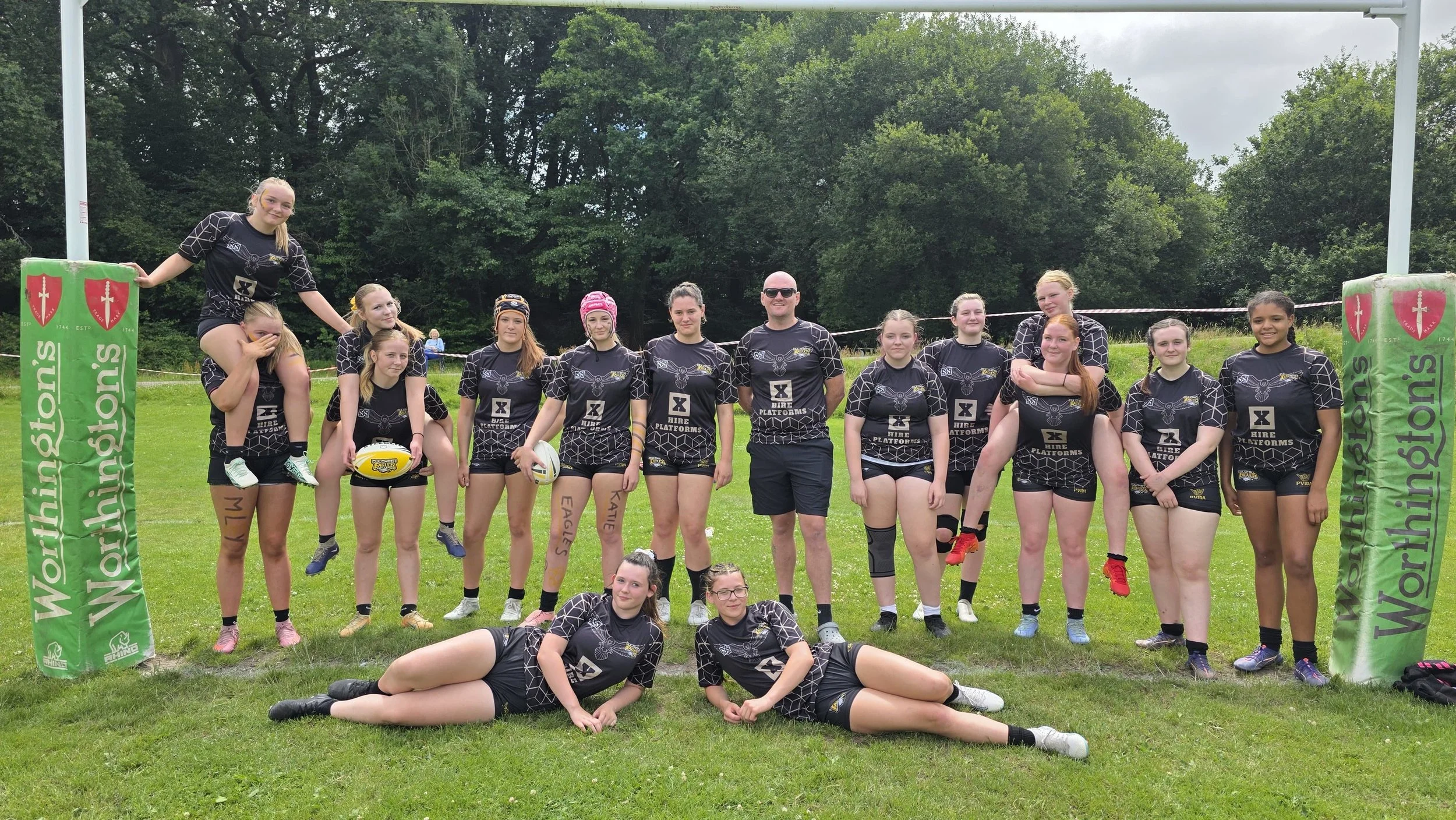 A group of young female rugby players and their coach posing for a team photo outdoors on a grassy field, with rugby posts and green trees in the background.