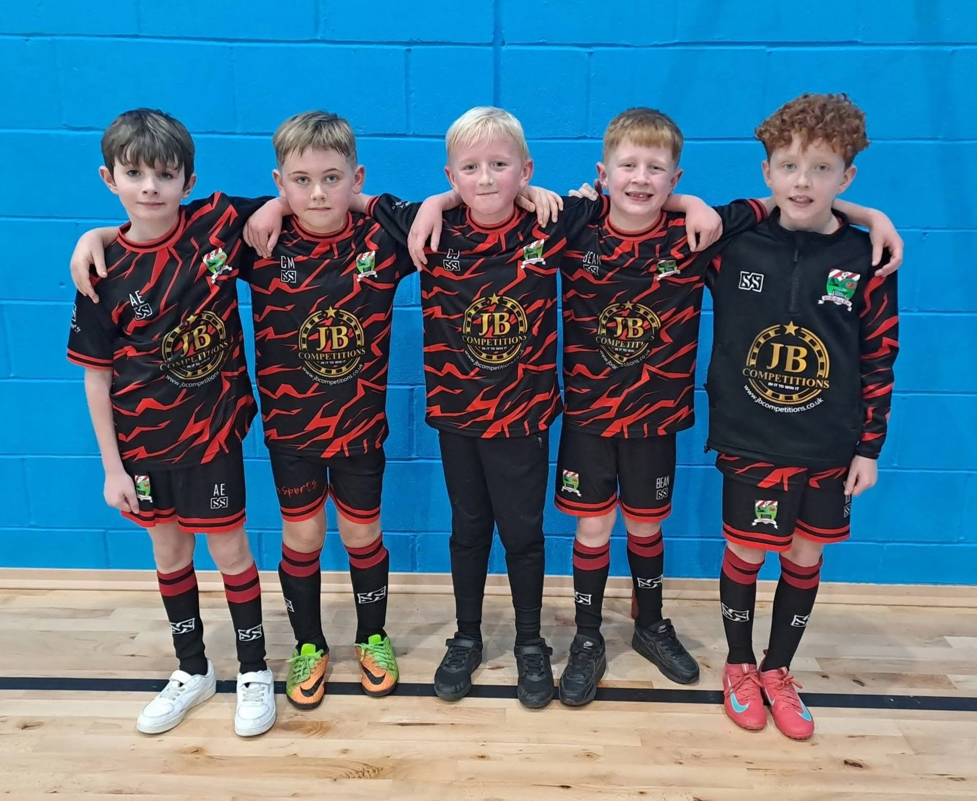 Group of five young boys standing against a blue wall, wearing matching black and red sports jerseys and black shorts, with their arms around each other's shoulders.