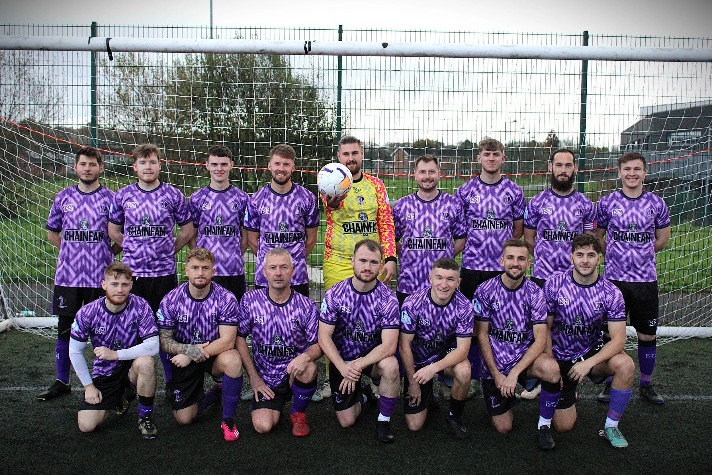 A group of young men in purple soccer uniforms with black shorts posing in front of a soccer goal on a field. One man in the middle wears a yellow goalie jersey holding a soccer ball. Everyone is smiling.