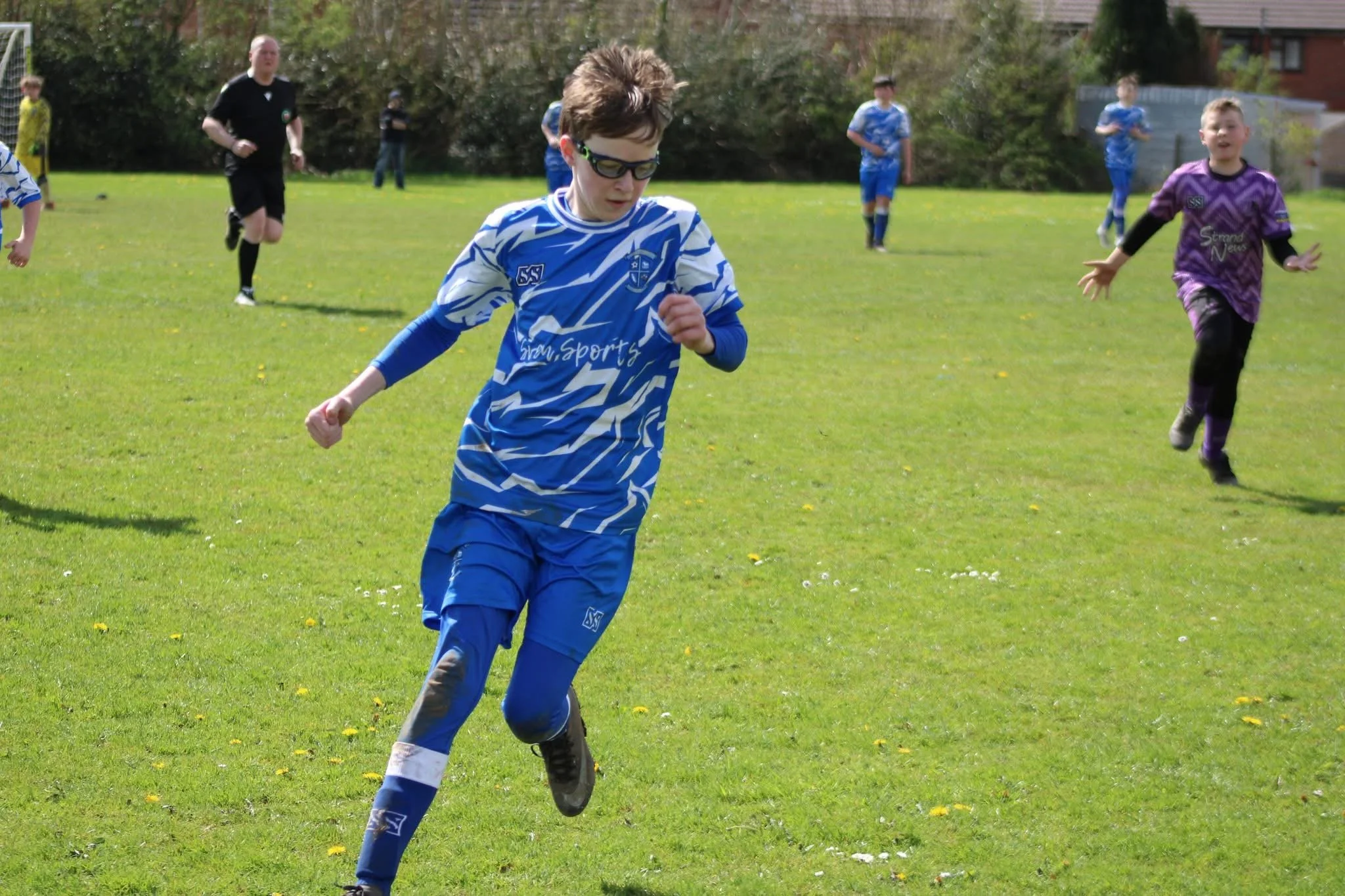 A young boy wearing sunglasses and a blue and white patterned soccer uniform running on a grassy soccer field during a game. Other children and an adult referee are in the background.