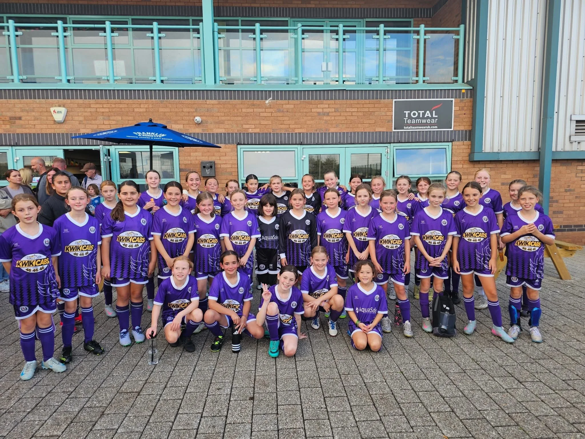 A large group of young female soccer players in purple uniforms posing for a group photo outside a building with a brick and metal exterior, some adults in the background, and a blue umbrella.