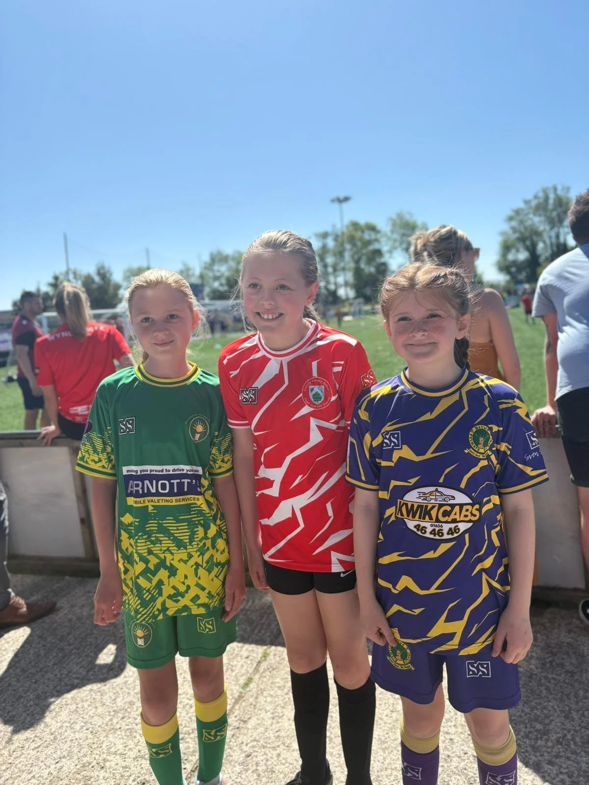 Three young girls in colorful soccer uniforms standing on a field with other players and spectators in the background, smiling at the camera. The girl on the left wears a green jersey, the middle girl wears a red jersey, and the girl on the right wears a blue and yellow jersey.