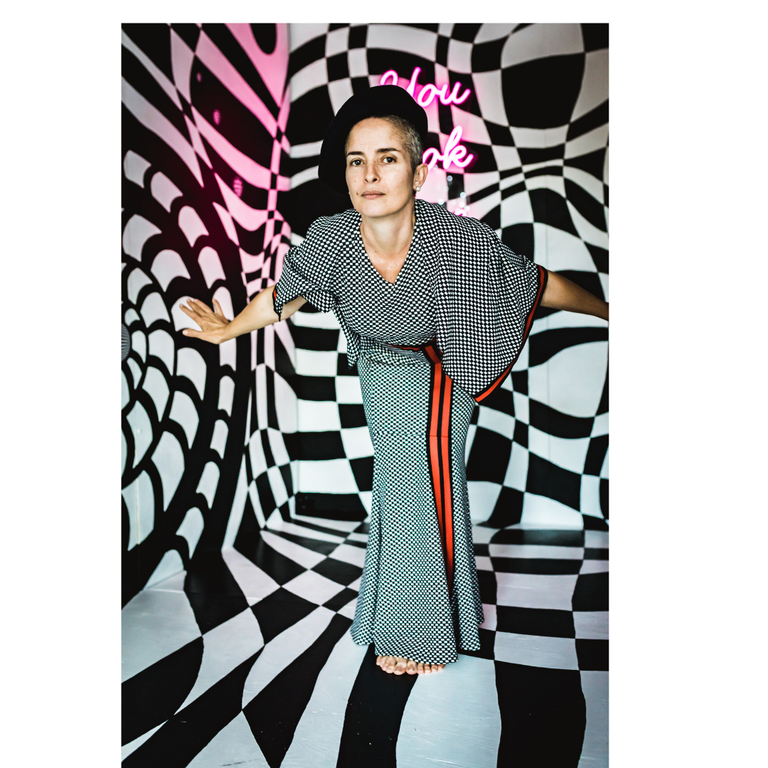 A woman dressed in black and white checkered clothing and wearing a black hat poses in front of a black and white optical illusion background with a pink neon sign that says "You look fine".