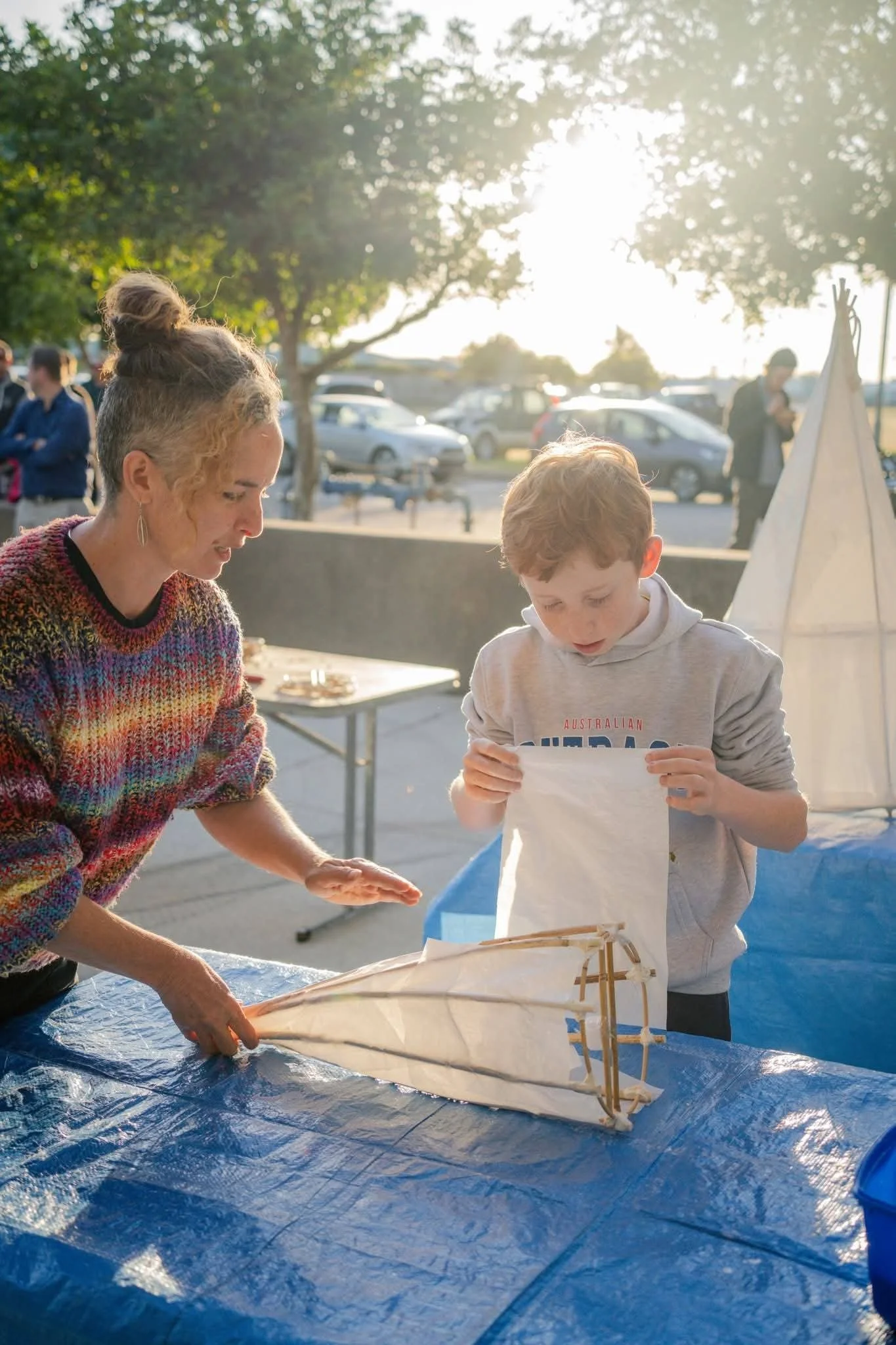 A woman and a boy assembling a small lantern on a blue table outdoors during late afternoon or early evening. The background shows other people, parked cars, trees, and bright sunlight.