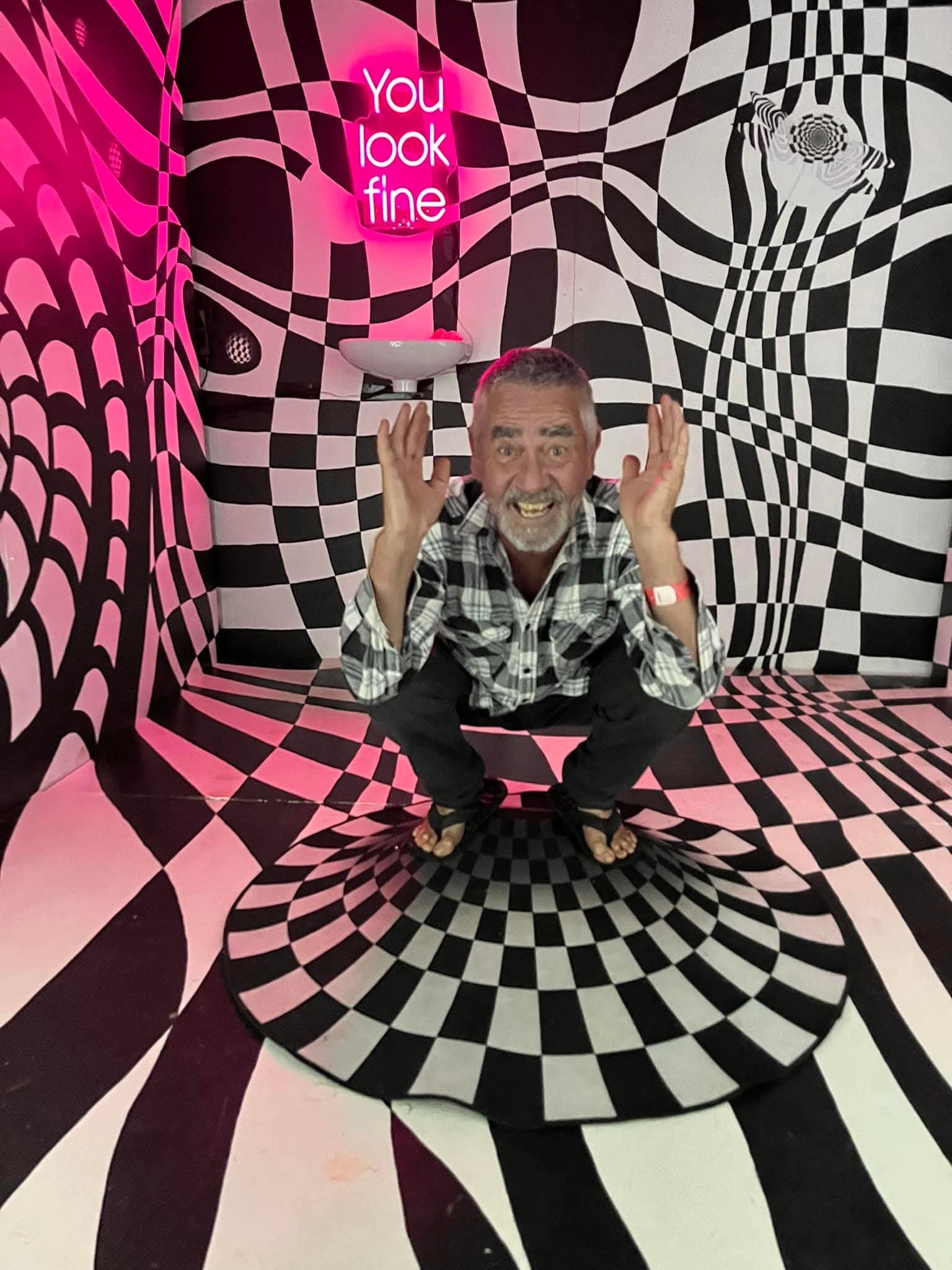 A man with gray hair and a beard, smiling and squatting, in a room with black and pink optical illusion wall and floor patterns. The wall has a neon sign that says 'You look fine.'