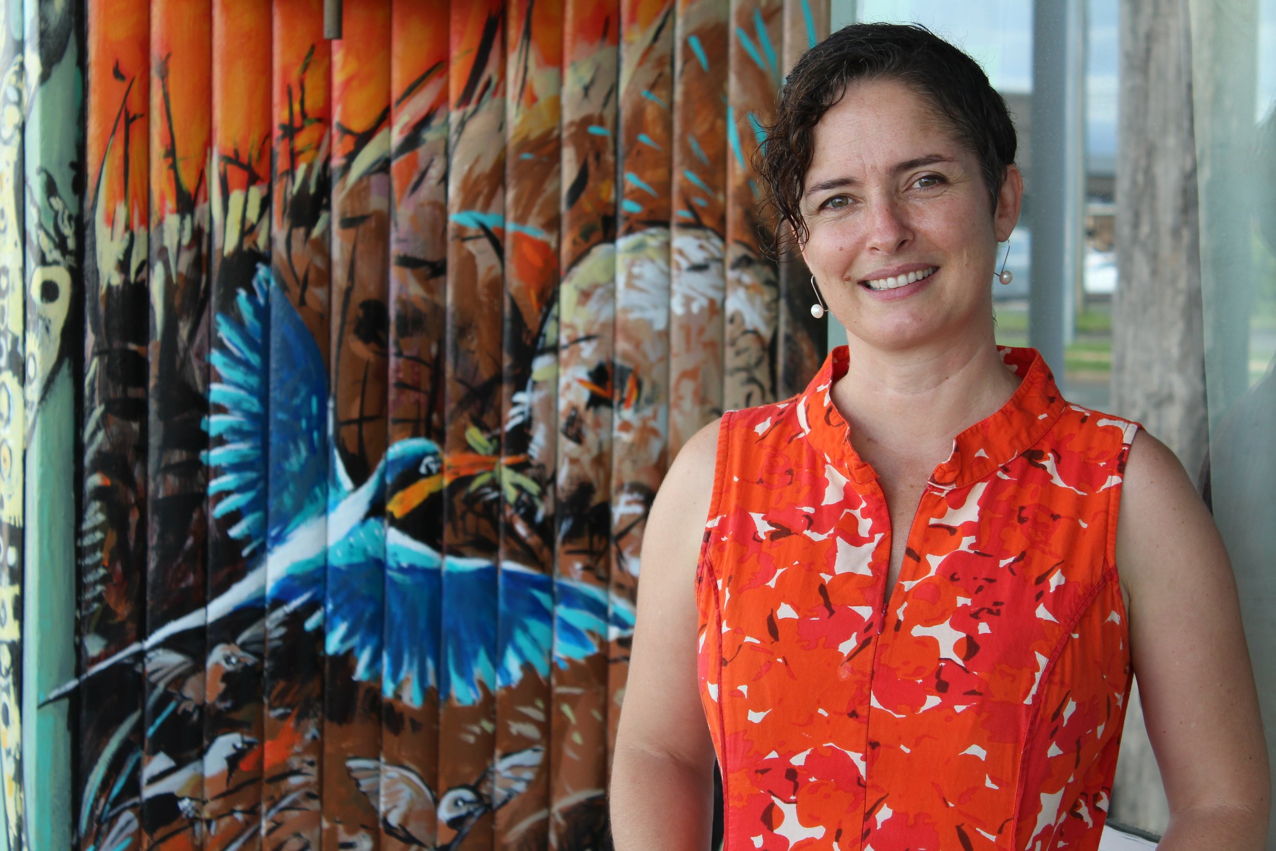 A woman with short, curly dark hair and pearl earrings smiling and standing in front of colorful street art painted on wooden panels.