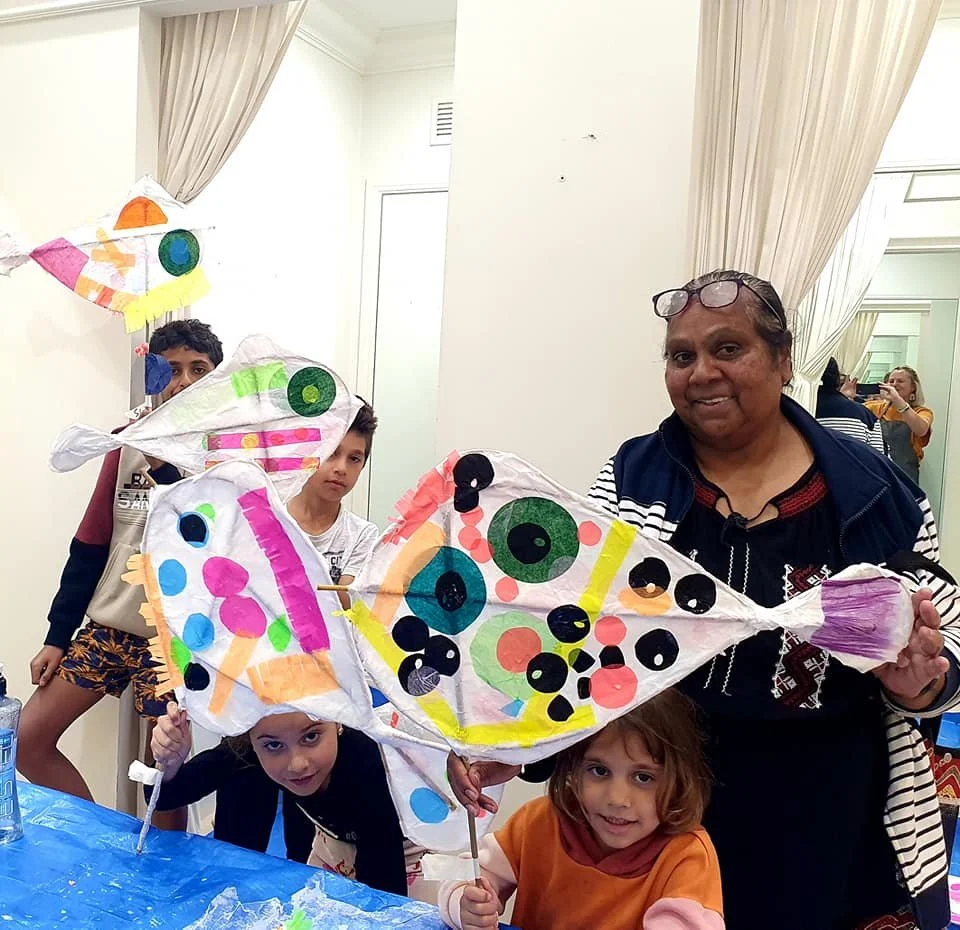 Children and an adult holding a large, colorful 2d sculpture decorated with circles and patterns, indoors with curtains and a mirror in the background.