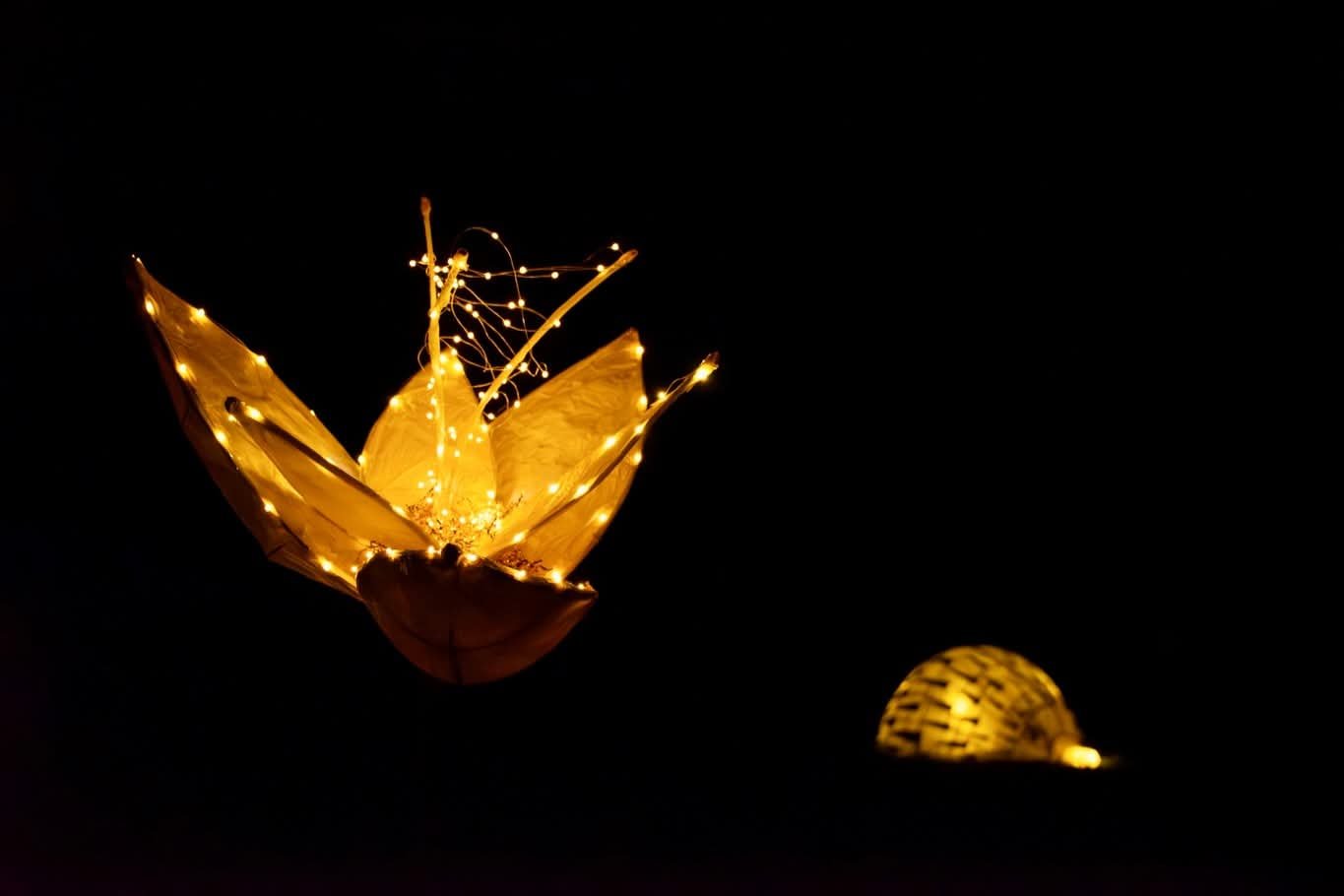 Decorative illuminated paper lantern shaped like a flower with fairy lights, and a smaller illuminated spherical object in the background, all against a dark background.
