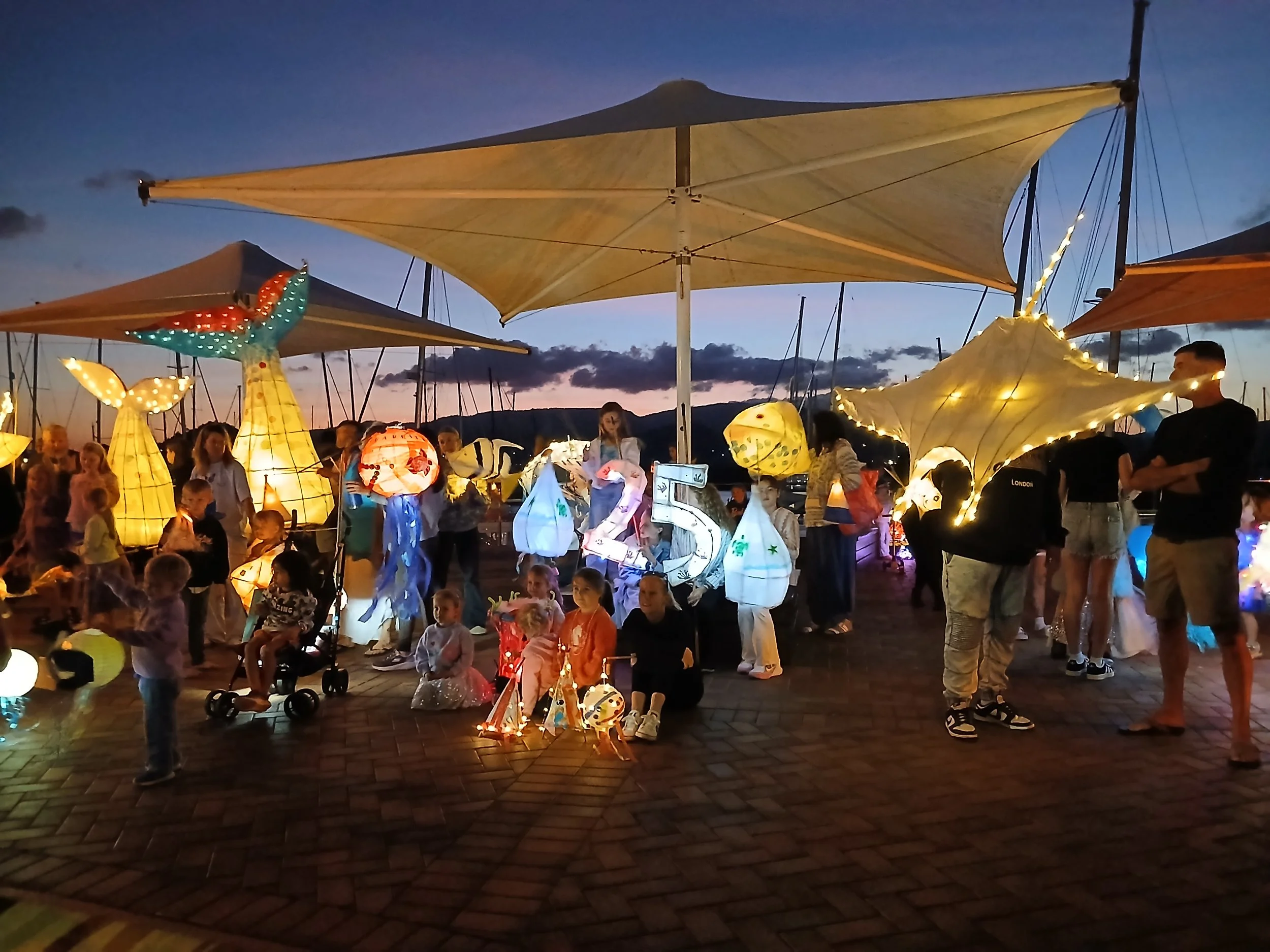 People gathered outdoors at dusk, holding illuminated lanterns and surrounded by decorated paper lanterns, with a marina and sailboats in the background.