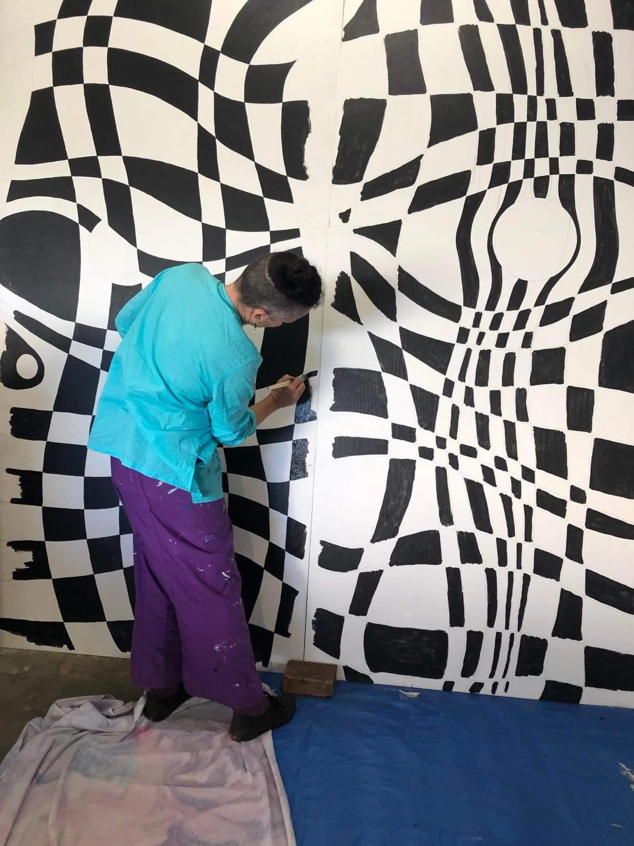 An artist creates a black and white geometric mural on a wall with a pattern resembling optical illusions, standing on a blue tarp and wearing purple pants and a turquoise shirt.