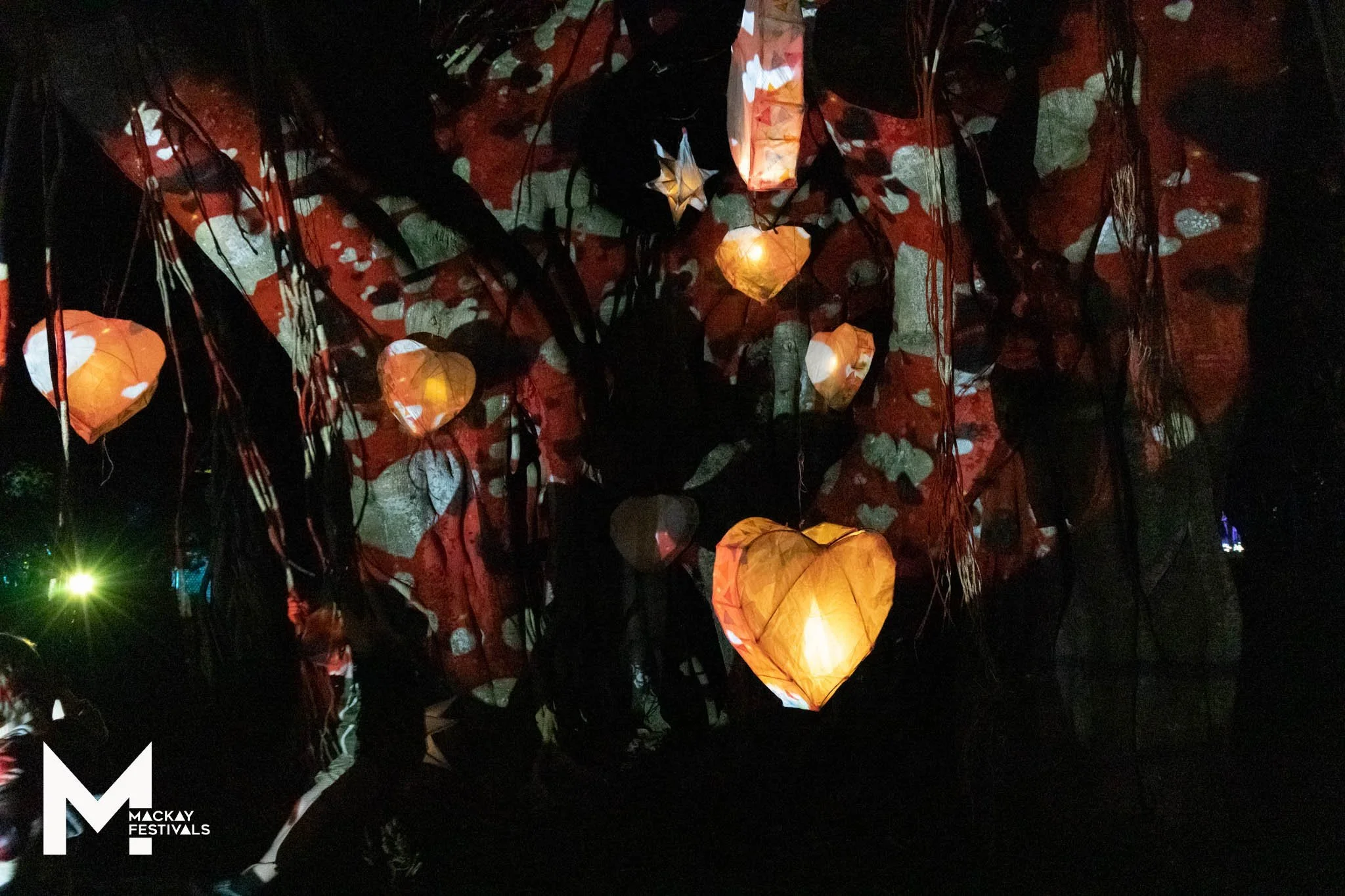 Night scene at a festival with illuminated paper lanterns hanging against a dark background with projections casting warm glow and shadows.