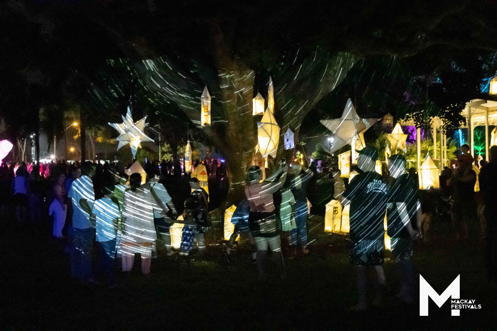 Night scene at a festival with illuminated lanterns hanging from trees and a crowd of people gathered underneath, with the Mackay Festivals logo in the bottom right corner.
