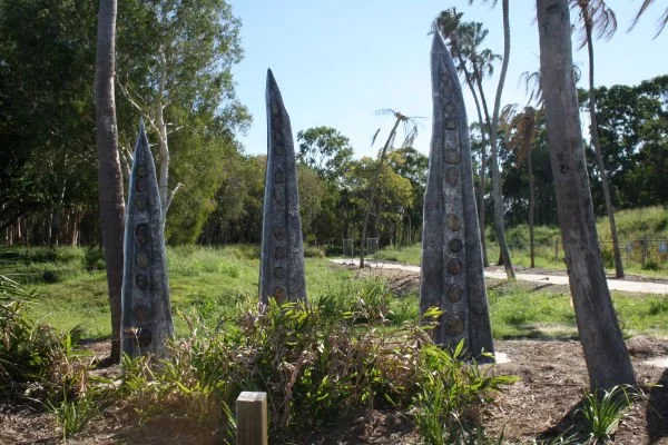 Four tall, slim, metal sculptures with ceramic tile inlays situated among green plants and trees in an outdoor setting.