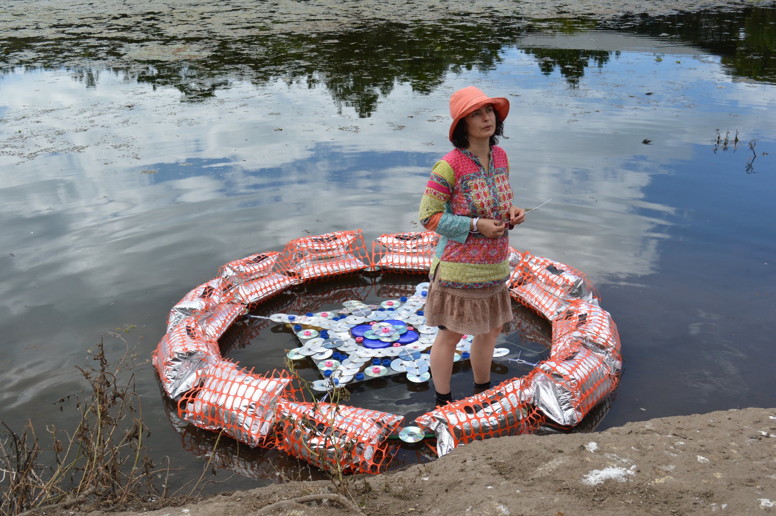 Woman standing in a floating sculpture made of plastic bottle caps and recycled materials, on a river, wearing a colorful dress and an orange hat, with trees and sky reflected in the water.