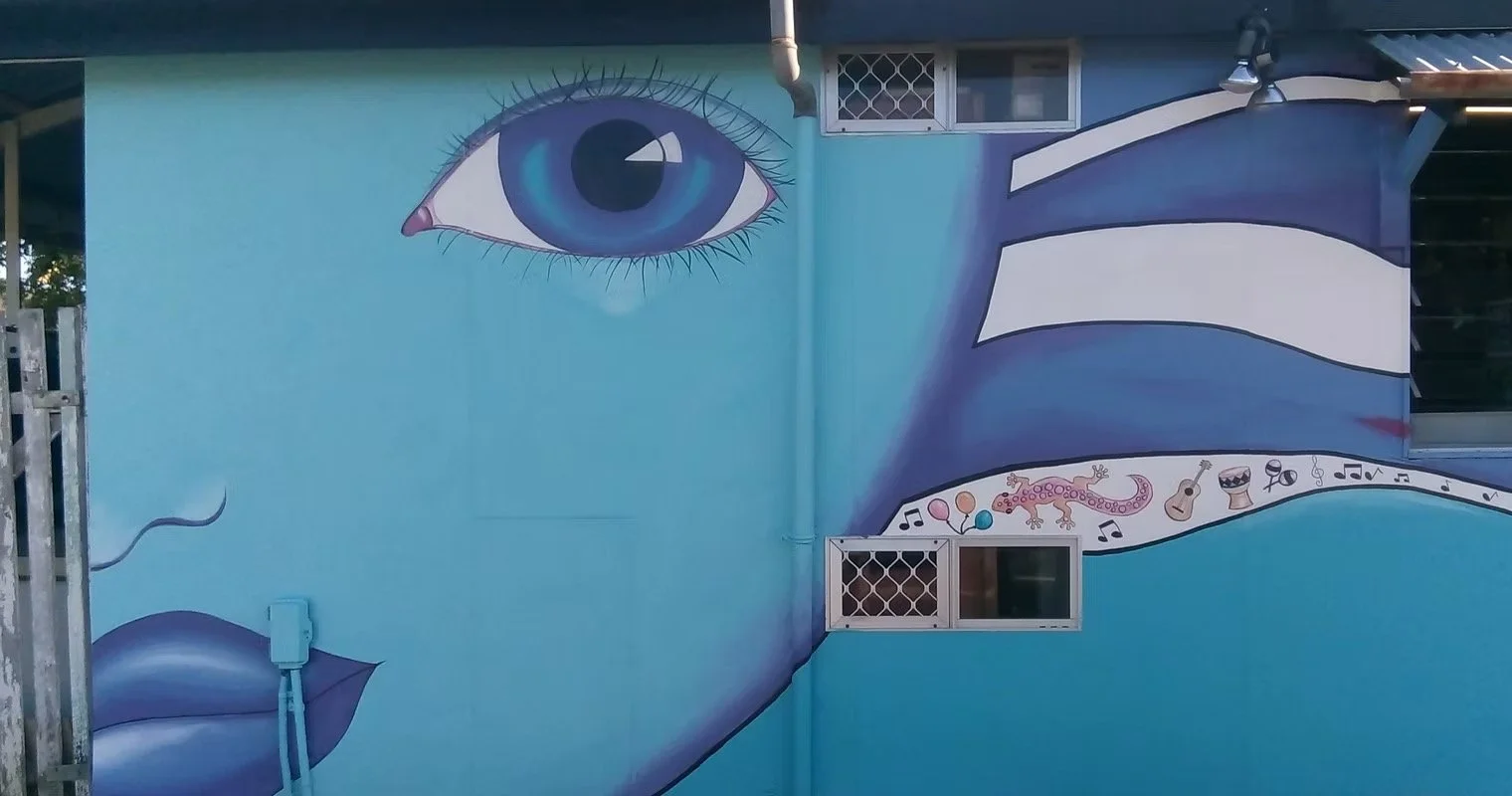 Colorful mural of a large eye with long eyelashes and a blue iris painted on the side of a building. The mural includes lips and a stream of musical symbols, a gecko, and various small illustrations like a guitar, drum, and balloons.