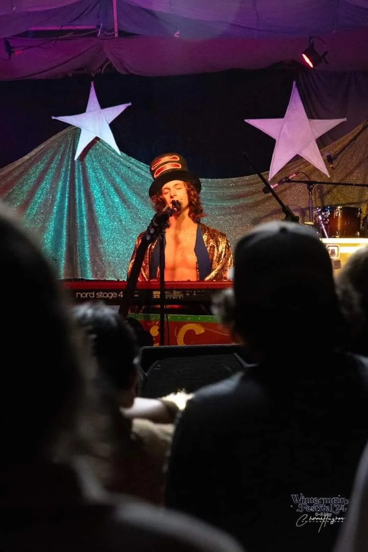 A musician dressed in a gold jacket and a striped top hat performs on stage at the Wintermoon Festival 24, with a colorful backdrop featuring two large stars designed by Lalune Croker Art.