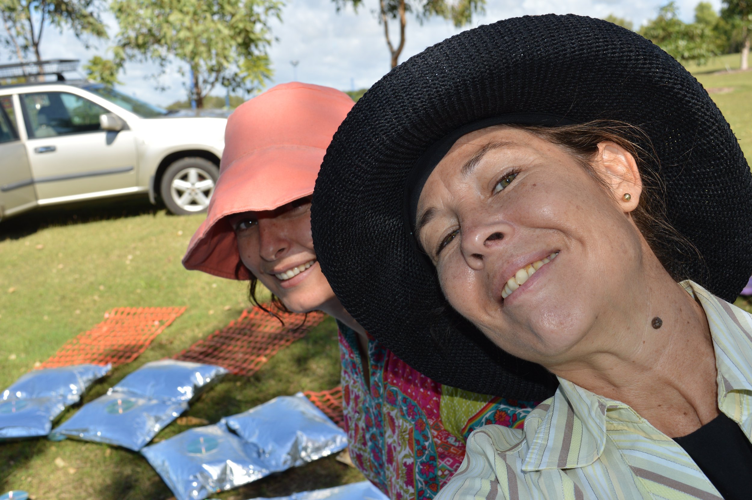 Two women smiling and wearing wide-brimmed hats outdoors, with colorful blankets and packages on the grass and a white car in the background.