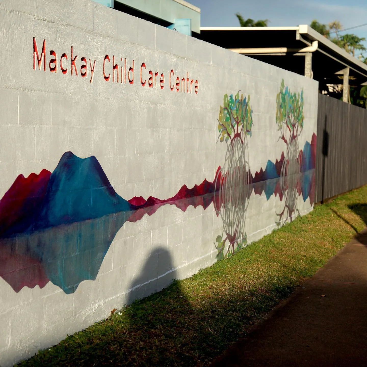 A colorful mural on a gray brick wall featuring mountains, trees, and a reflection of the landscape, with the text 'Mackay Child Care Centre' in red letters at the top left.