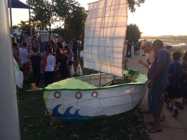 A small boat-shaped lantern float with a sail, decorated with waves, on display at an outdoor event during sunset, with people gathered around.