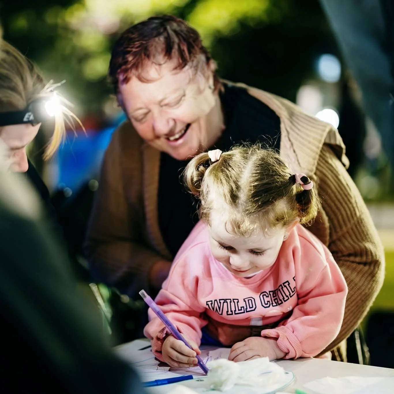 A young girl with two pigtails and a pink sweatshirt is working on her lantern puppet. An elderly woman is smiling and helping her during the workshop.