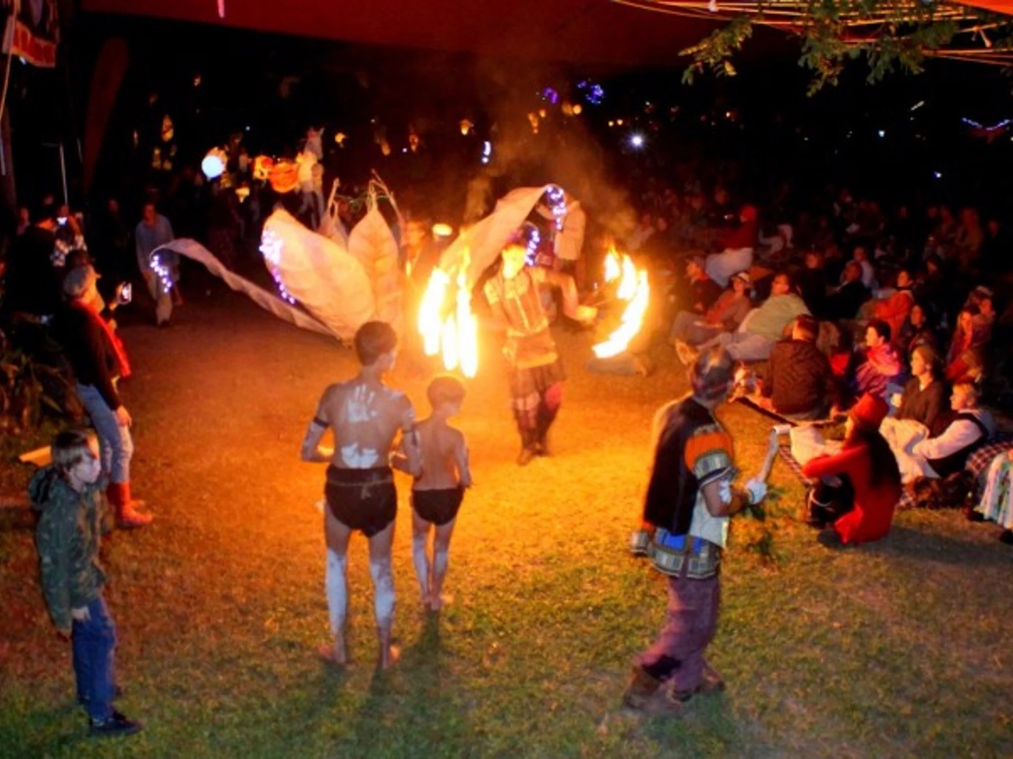 A nighttime outdoor gathering featuring a large lantern, a fire dance performance and an indigenous team with a crowd of people watching.