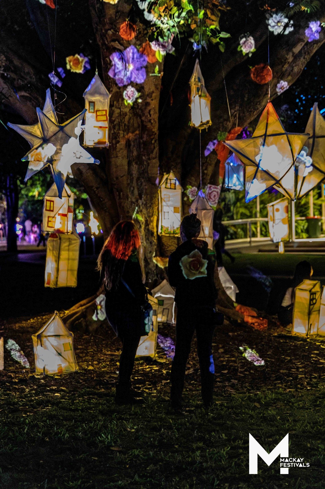 People walking under decorative paper lanterns shaped like stars and houses hanging from a tree at night during a festival, with flowers and lights in the background.