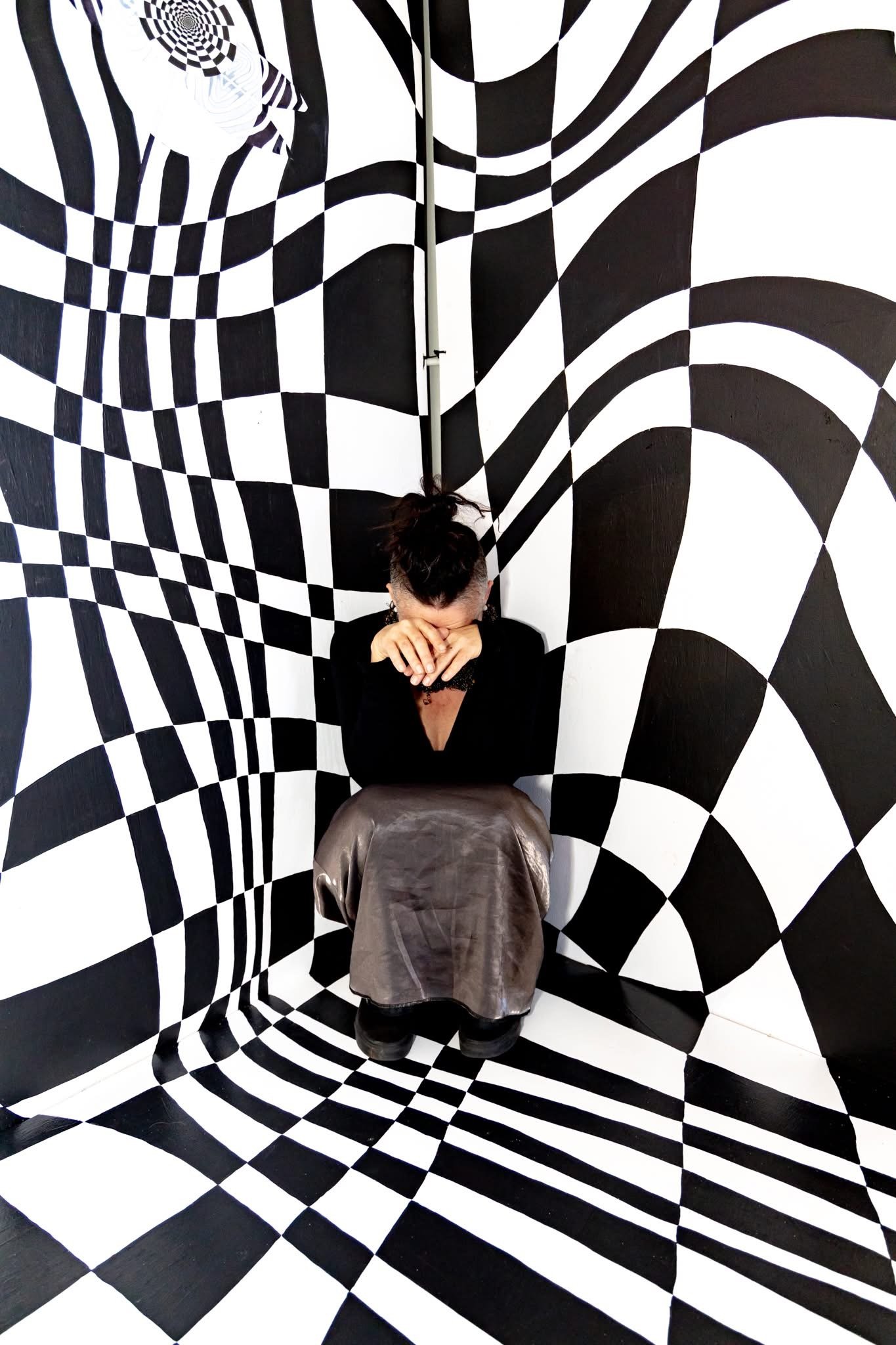 A person sitting with their hands covering their face in front of a black and white optical illusion wall pattern that gives the appearance of a tunnel or vortex.