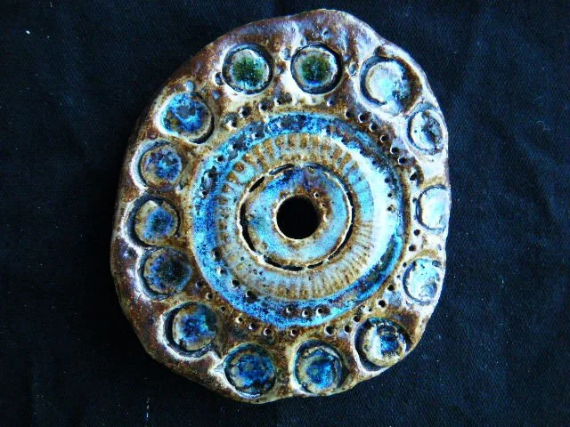 A round, ceramic art piece with concentric circles and a hole in the center, decorated with multiple small circular indentations filled with blue, green, and white glazes.