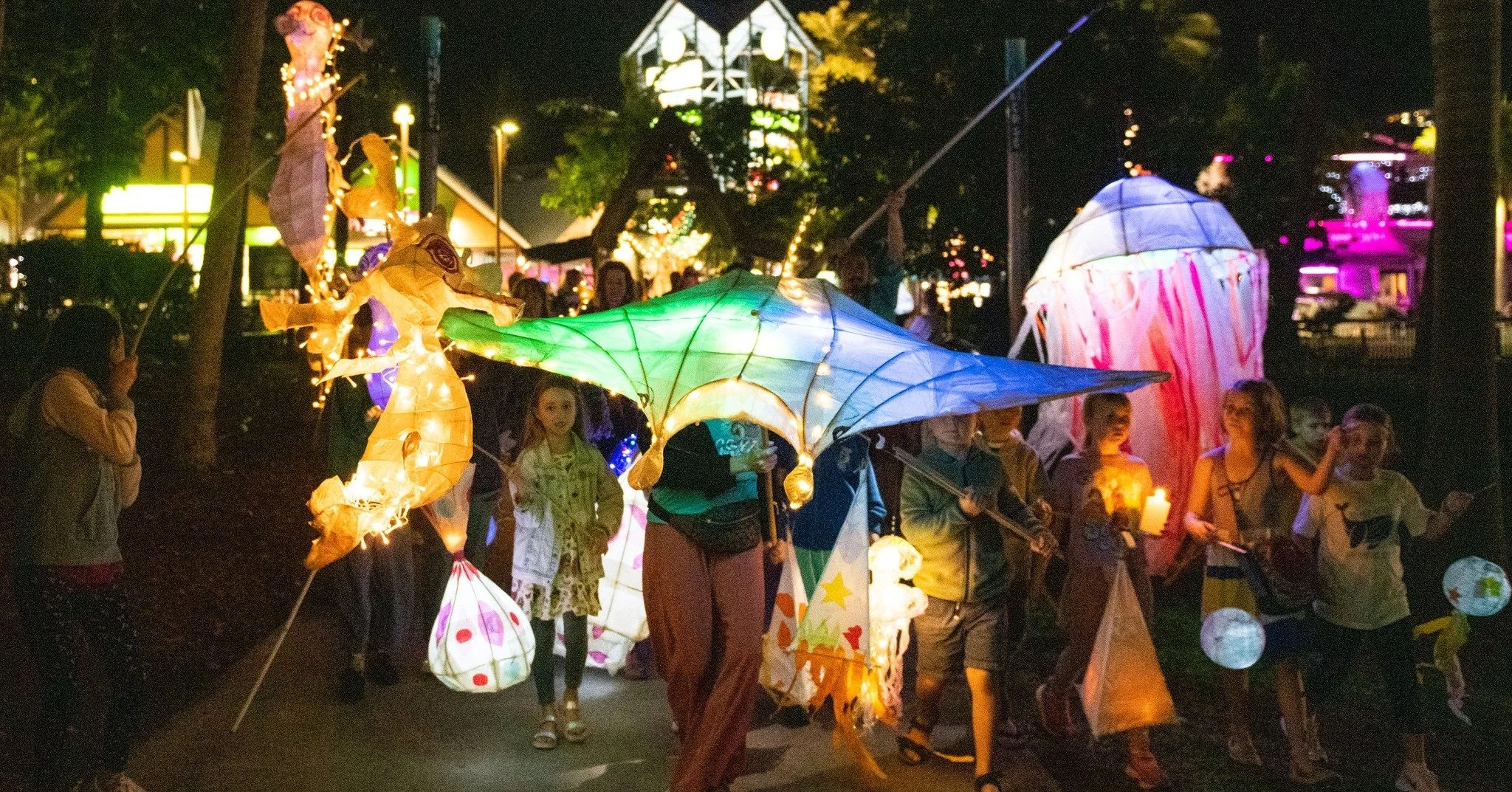 People participating in a nighttime lantern festival, holding colorful illuminated paper lanterns, surrounded by trees and buildings.