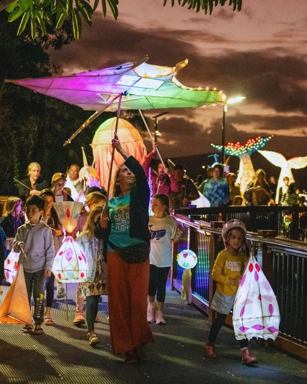 Children and adults walking at night in a lantern parade, holding colorful illuminated paper lanterns with a dark, cloudy sky in the background.